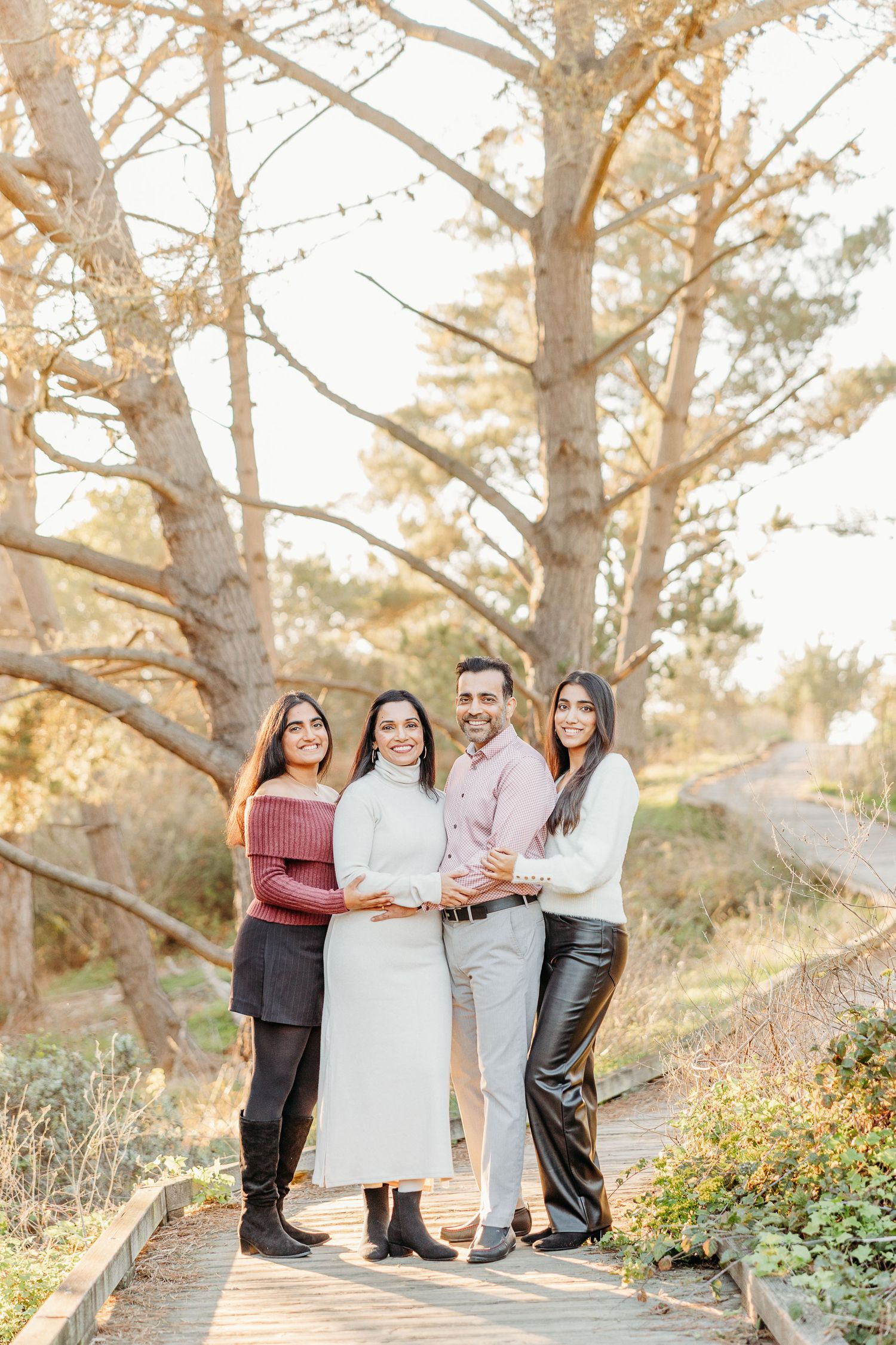 Family group in coordinated white outfits poses together among tall trees at sunset.