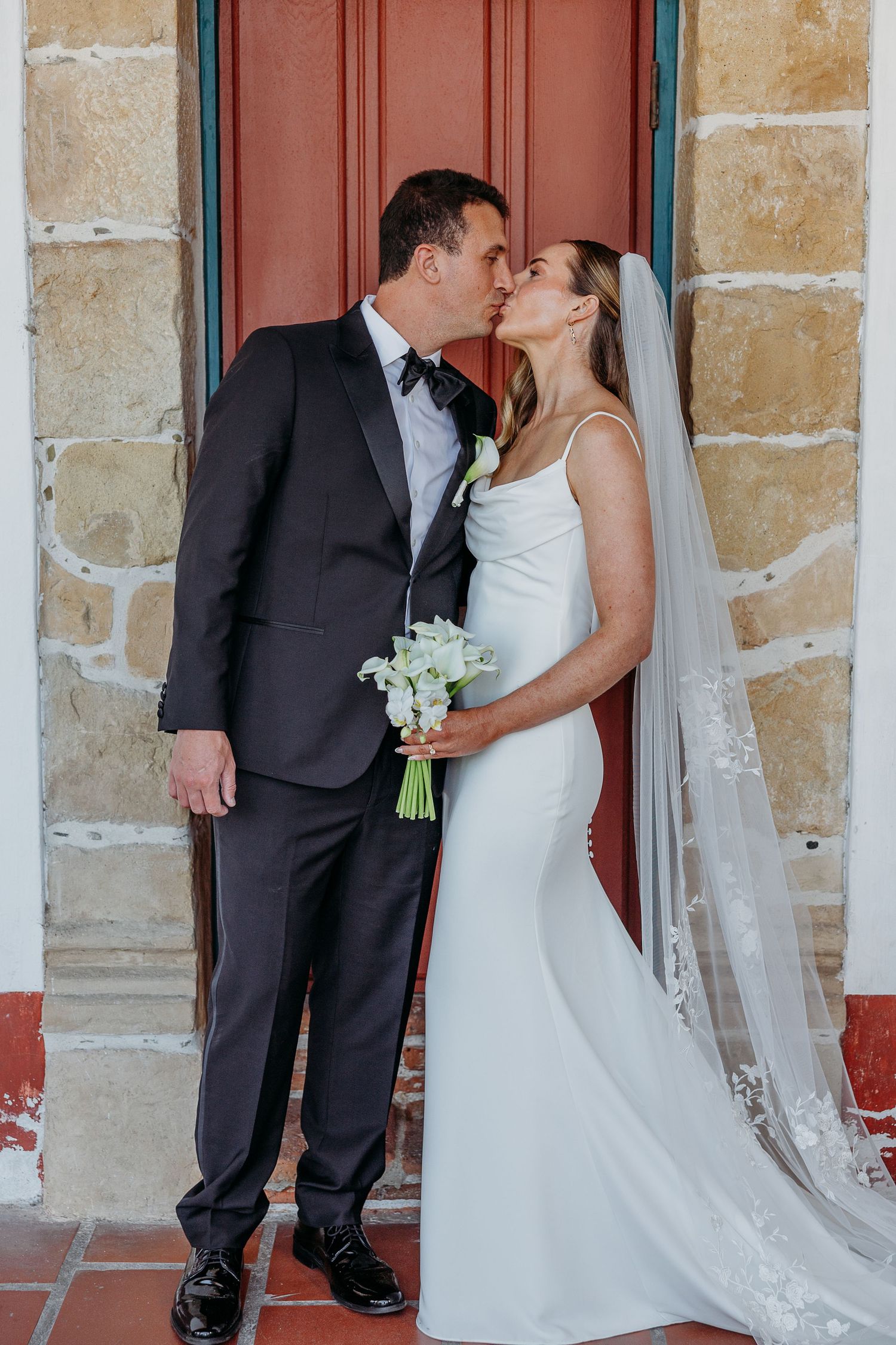 A romantic wedding couple sharing a kiss in front of a rustic red door with stone walls.