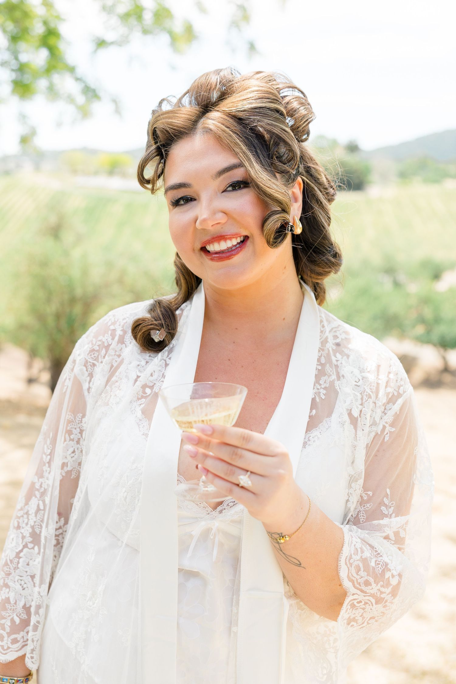 Smiling bride in white lace robe holding champagne glass outdoors at vineyard wedding.
