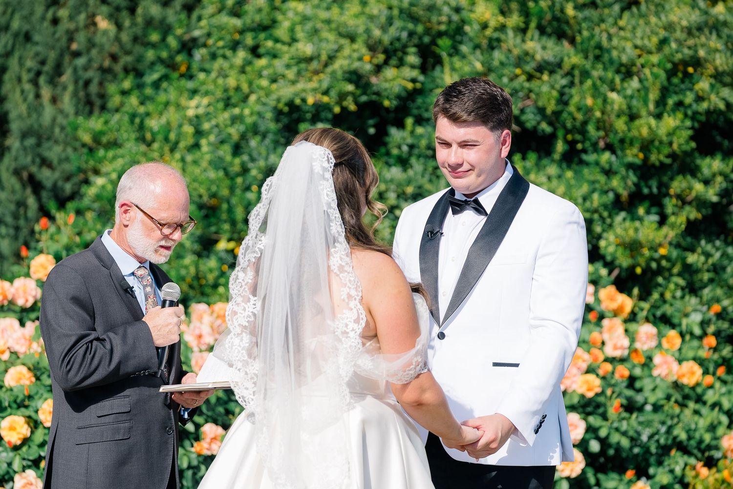 Groom in white jacket exchanges vows with bride before officiant surrounded by yellow roses at ceremony.