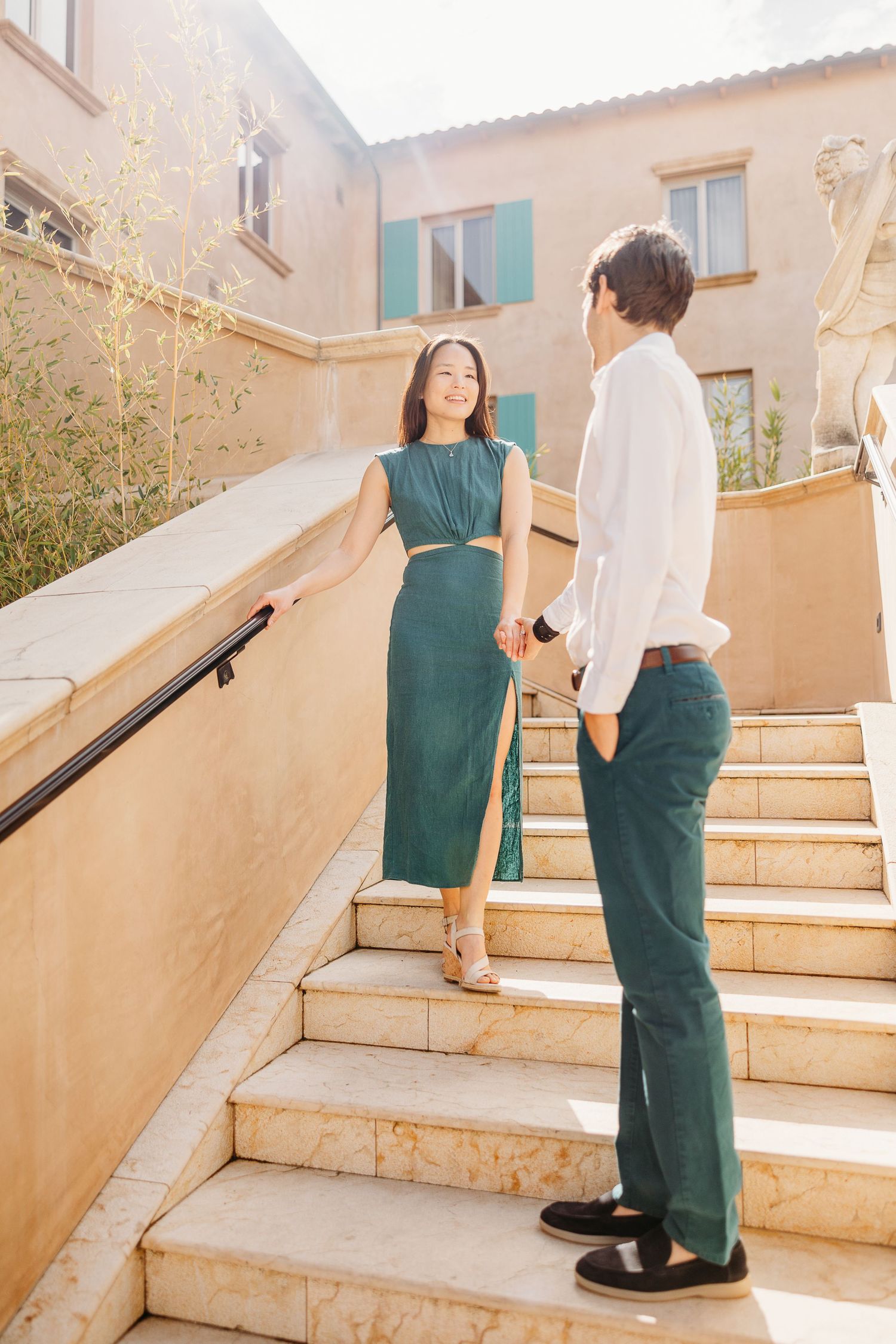 Two people in contrasting outfits walking up stone steps in warm sunlight against a beige building exterior.
