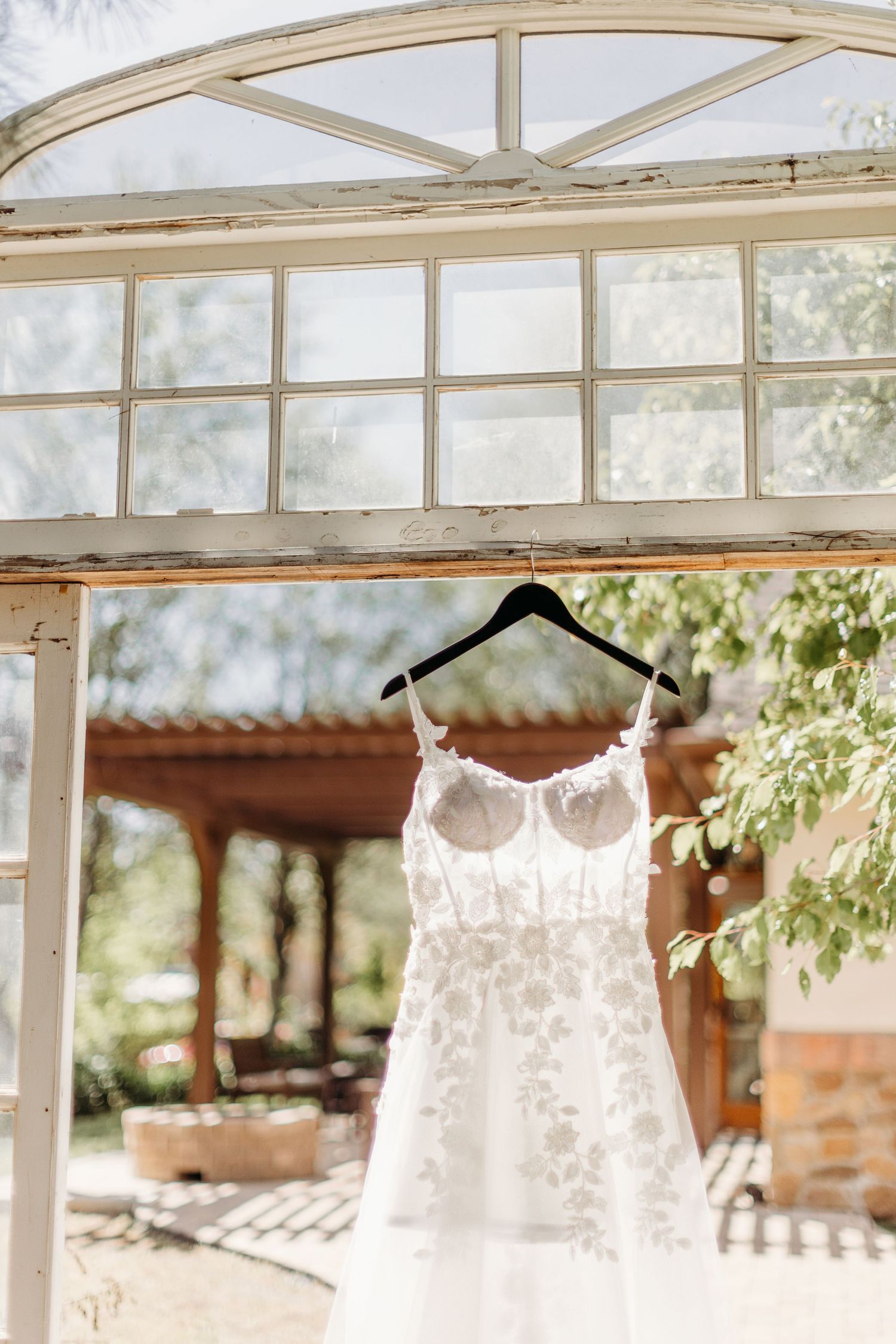 A delicate white lace wedding dress hanging from a hanger against an arched window frame in natural light.