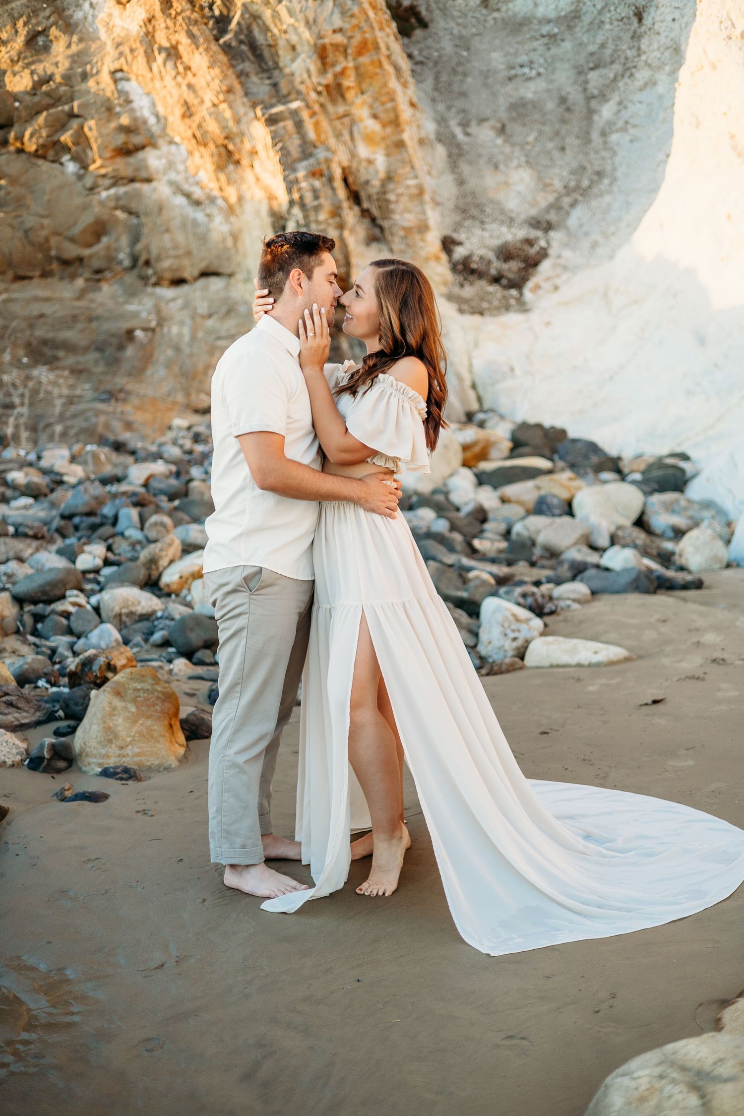A romantic couple embraces on beach with flowing white dress and coastal cliffs.