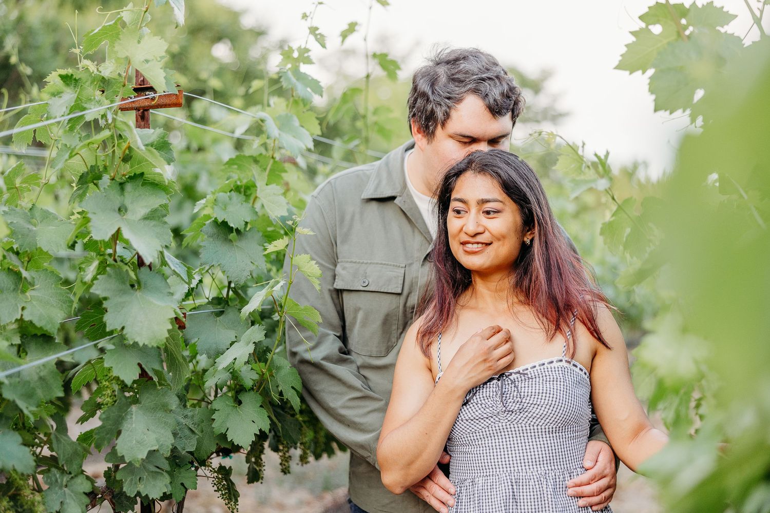 A couple smiles while posing together in a lush green vineyard on a sunny day.