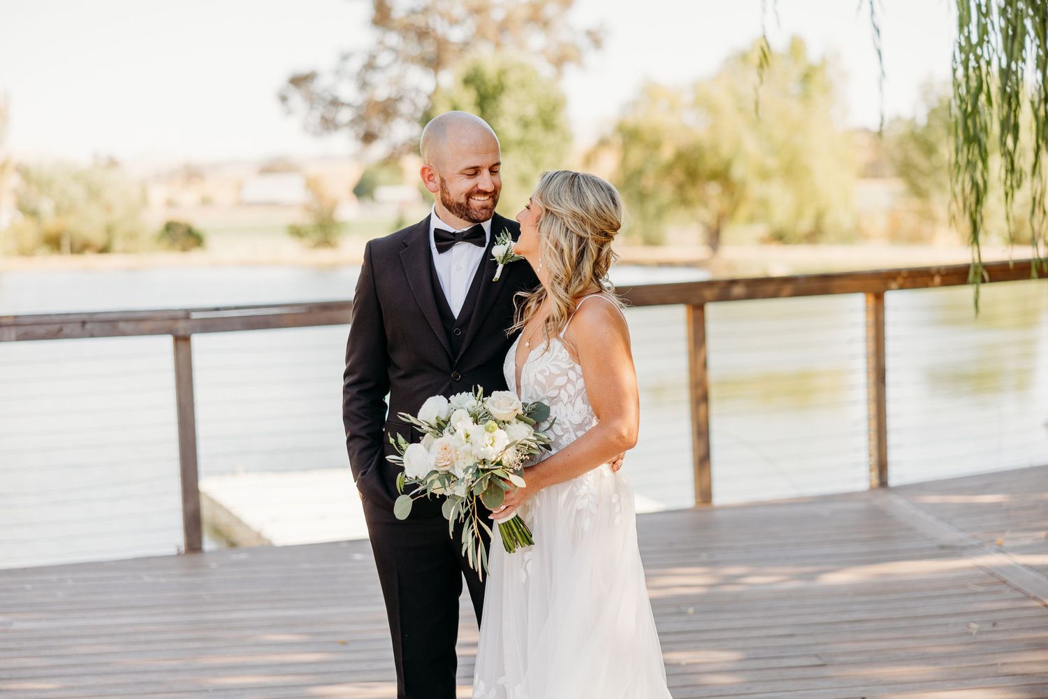 Bride holding white bouquet stands with groom on wooden deck near water.