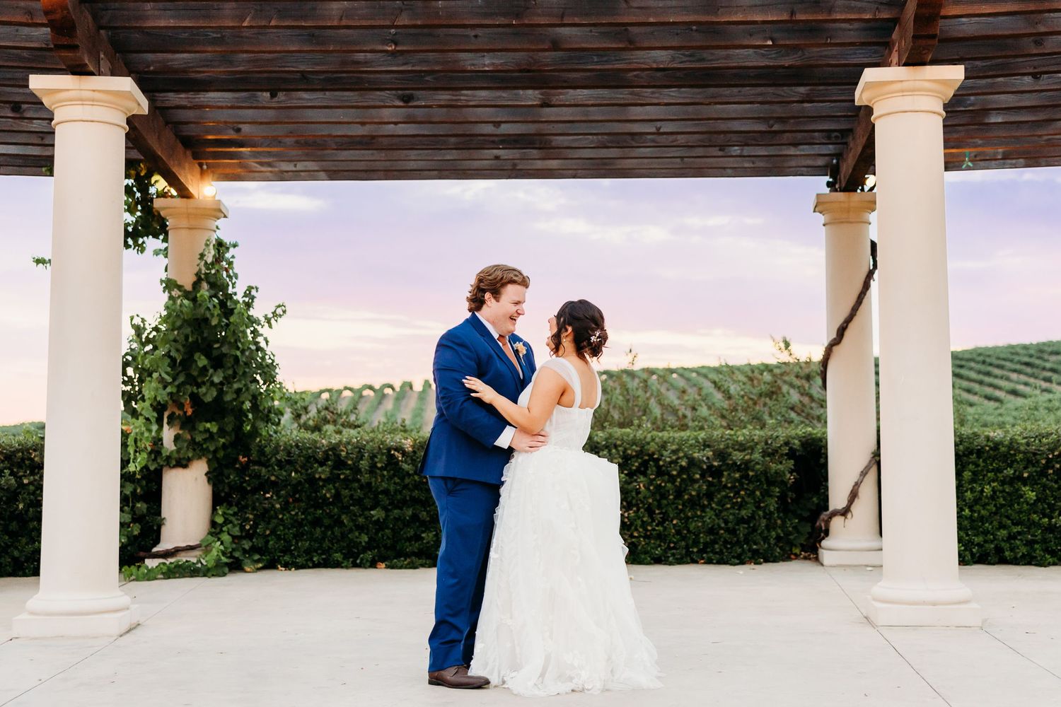 A couple dances together beneath a wooden pergola at a scenic winery venue.
