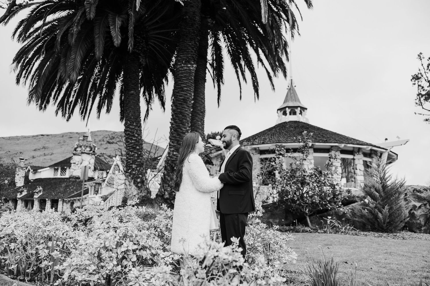 A couple in wedding attire embrace in a garden setting with a palm tree and gazebo in black and white photography.
