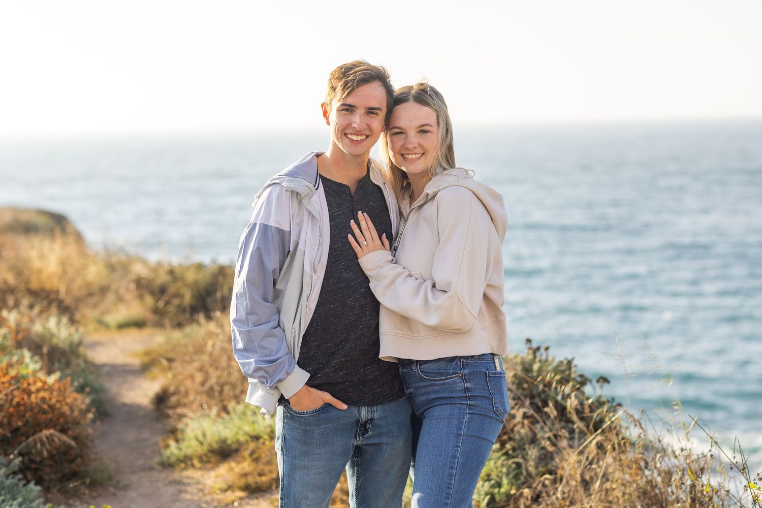 A couple embraces while standing on a coastal cliff overlooking the ocean on a sunny day.