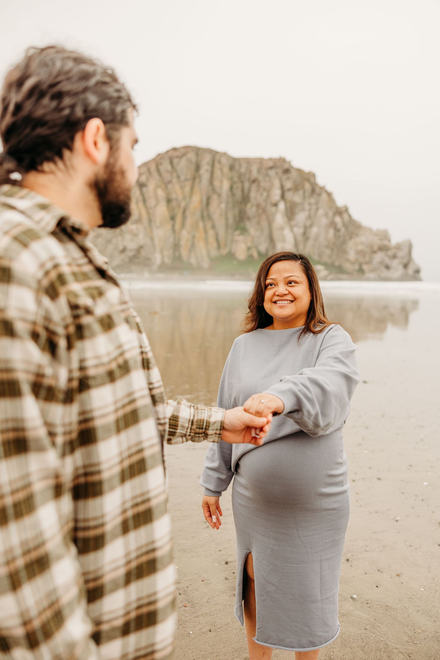 A couple holds hands on a beach with a large rock formation visible in the misty background.