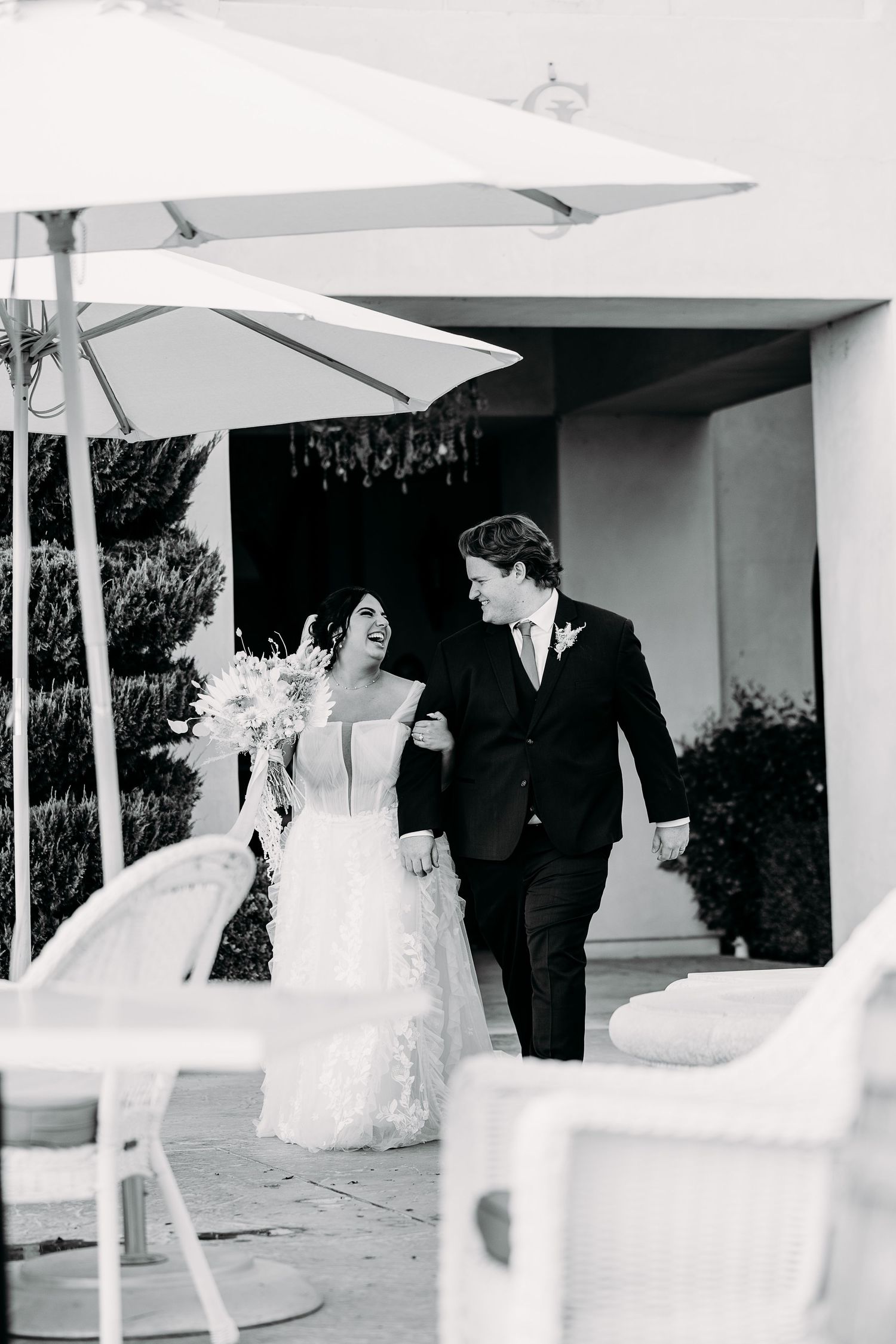 Black and white photo of a wedding couple walking together under white umbrellas on a patio.