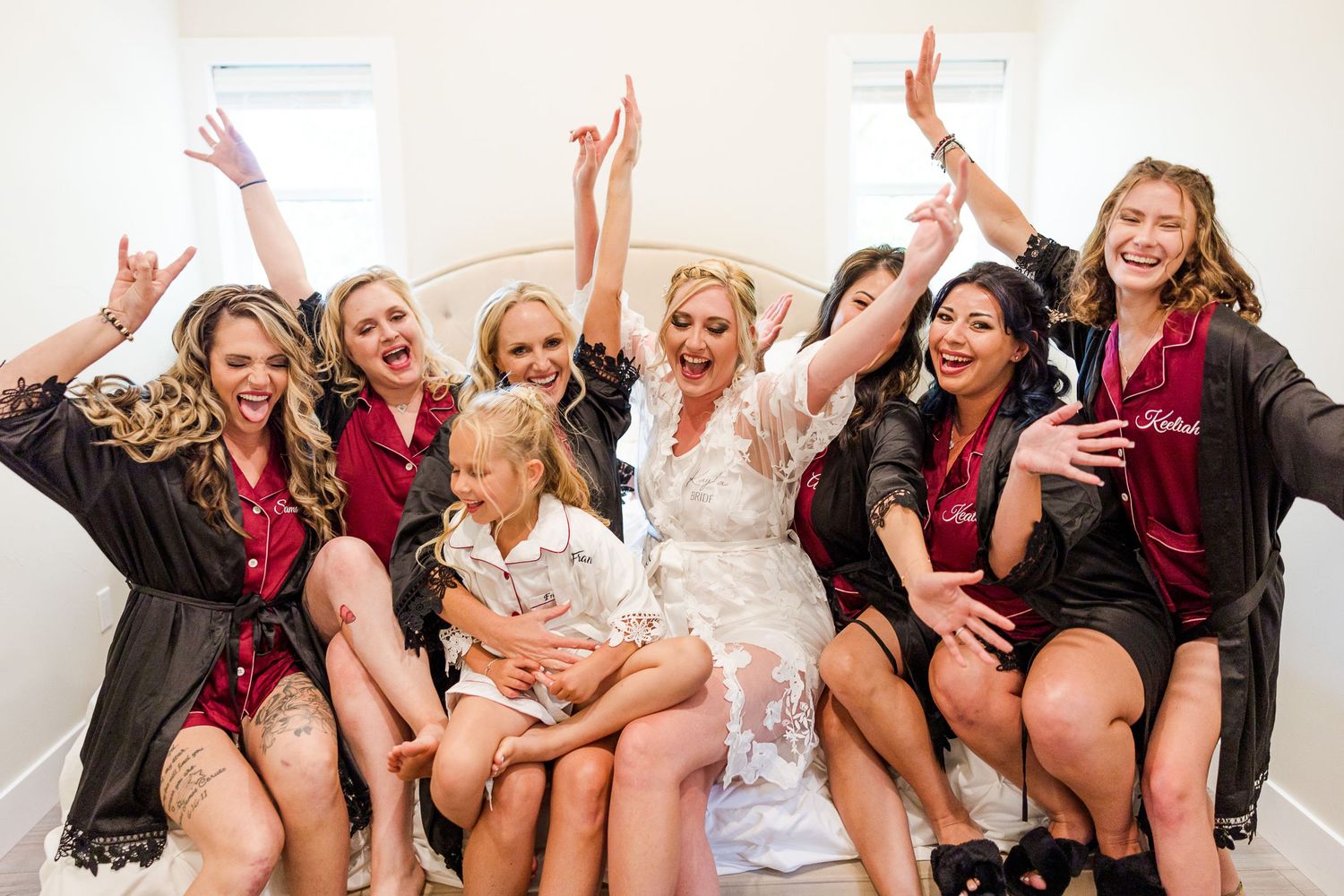 Wedding party celebrates joyfully in matching robes while sitting on white couch during pre-ceremony preparations.