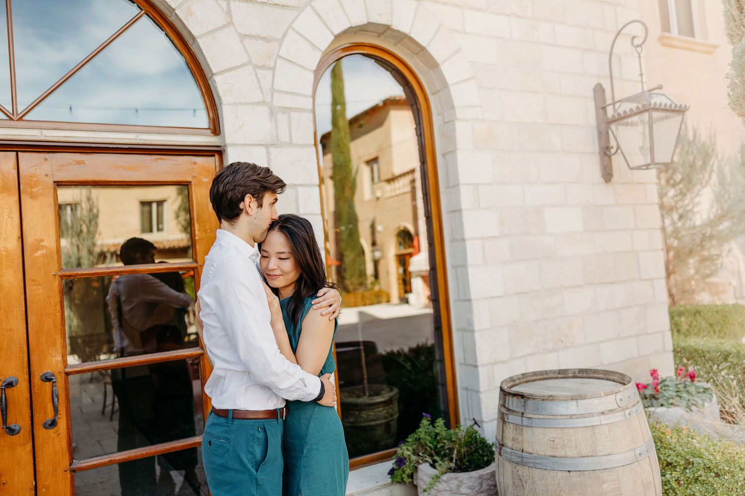 A couple embraces by rustic wooden doors and stone archways beside a wine barrel.