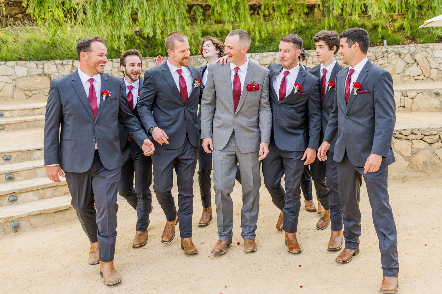Wedding party in gray and navy suits with burgundy ties laugh together on outdoor stone steps.