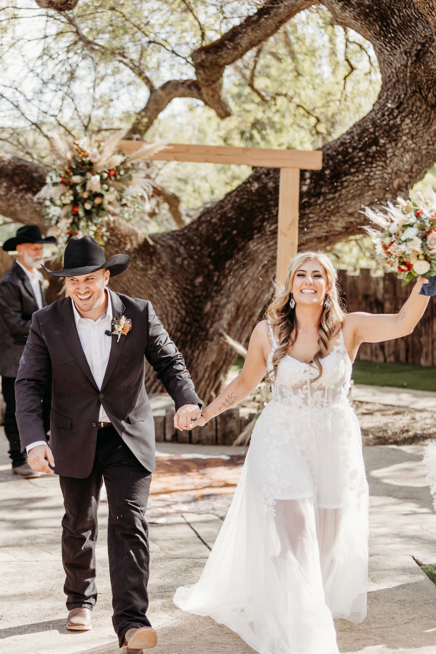 Joyful newlyweds walk hand in hand under a large tree at an outdoor wedding ceremony.