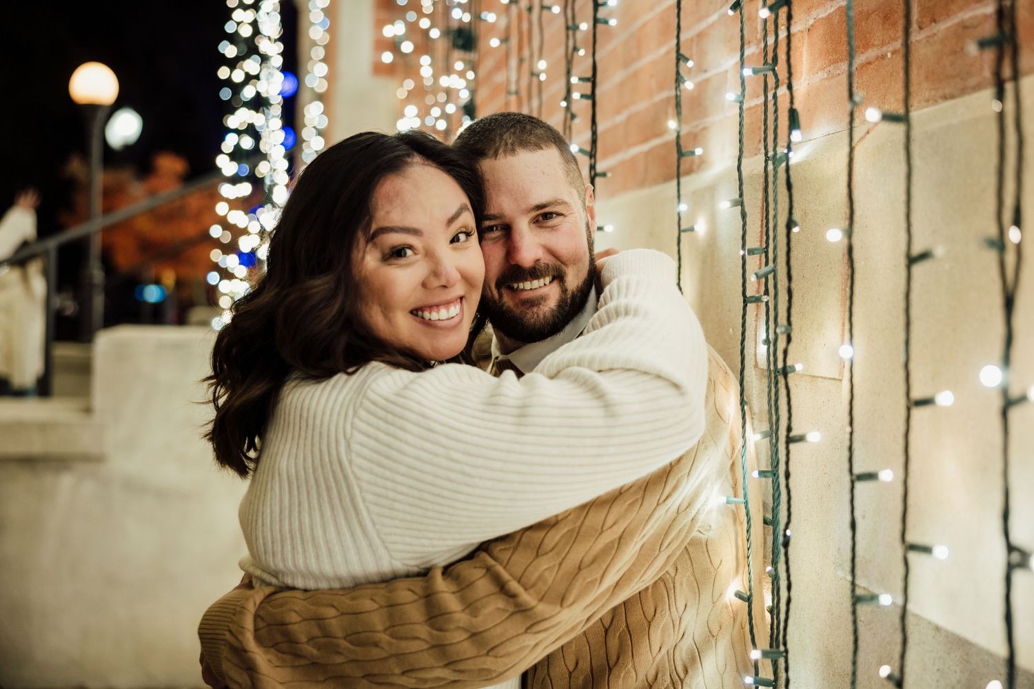 A couple embraces warmly under glowing string lights creating a romantic holiday atmosphere.