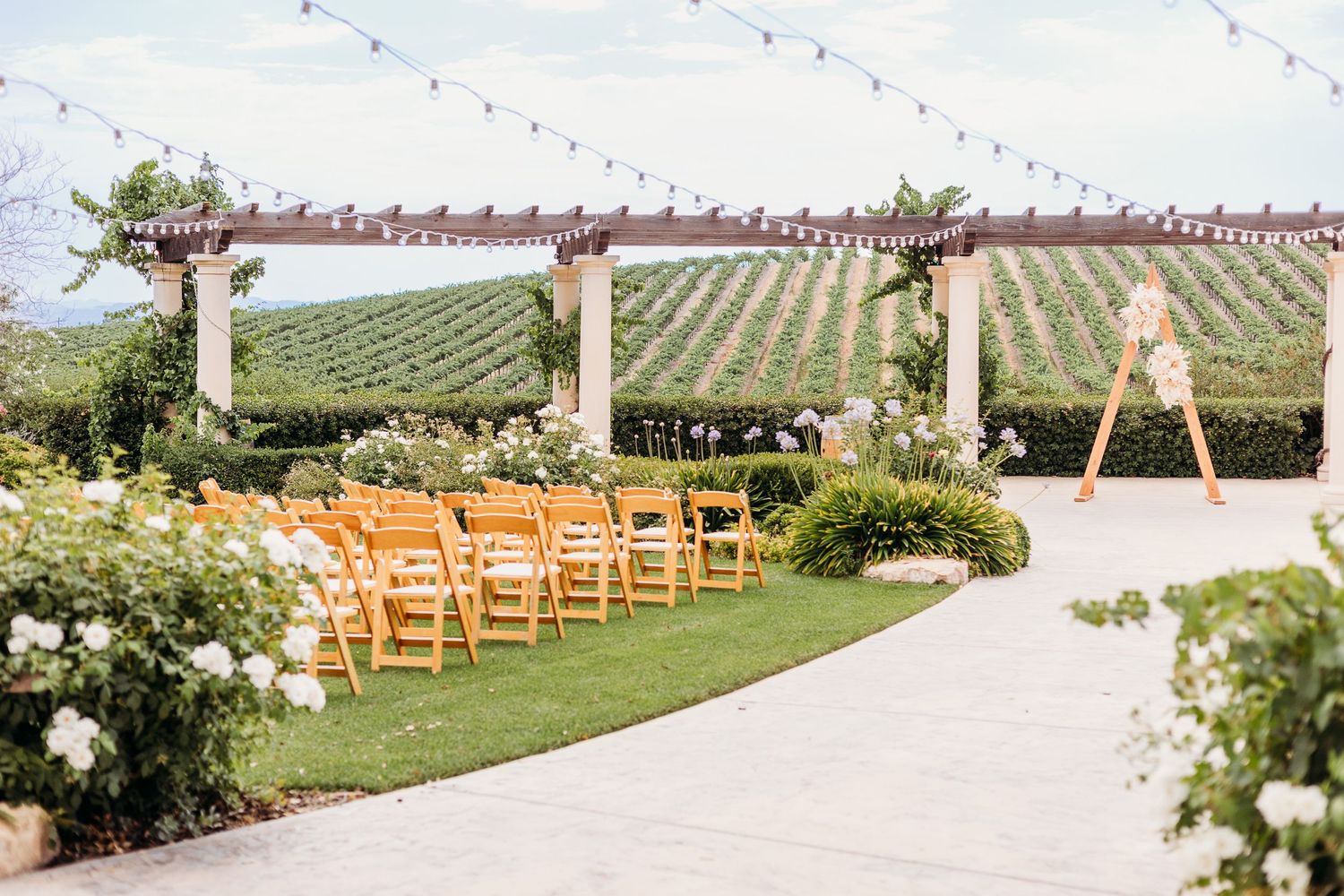 Outdoor wedding ceremony setup with wooden pergola and rows of chairs overlooking a vineyard.