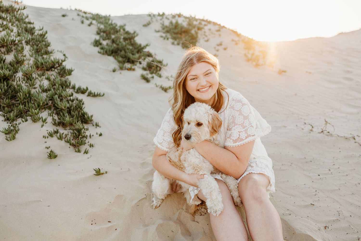 Graduate in white lace dress cuddles small white puppy on sandy beach dunes.