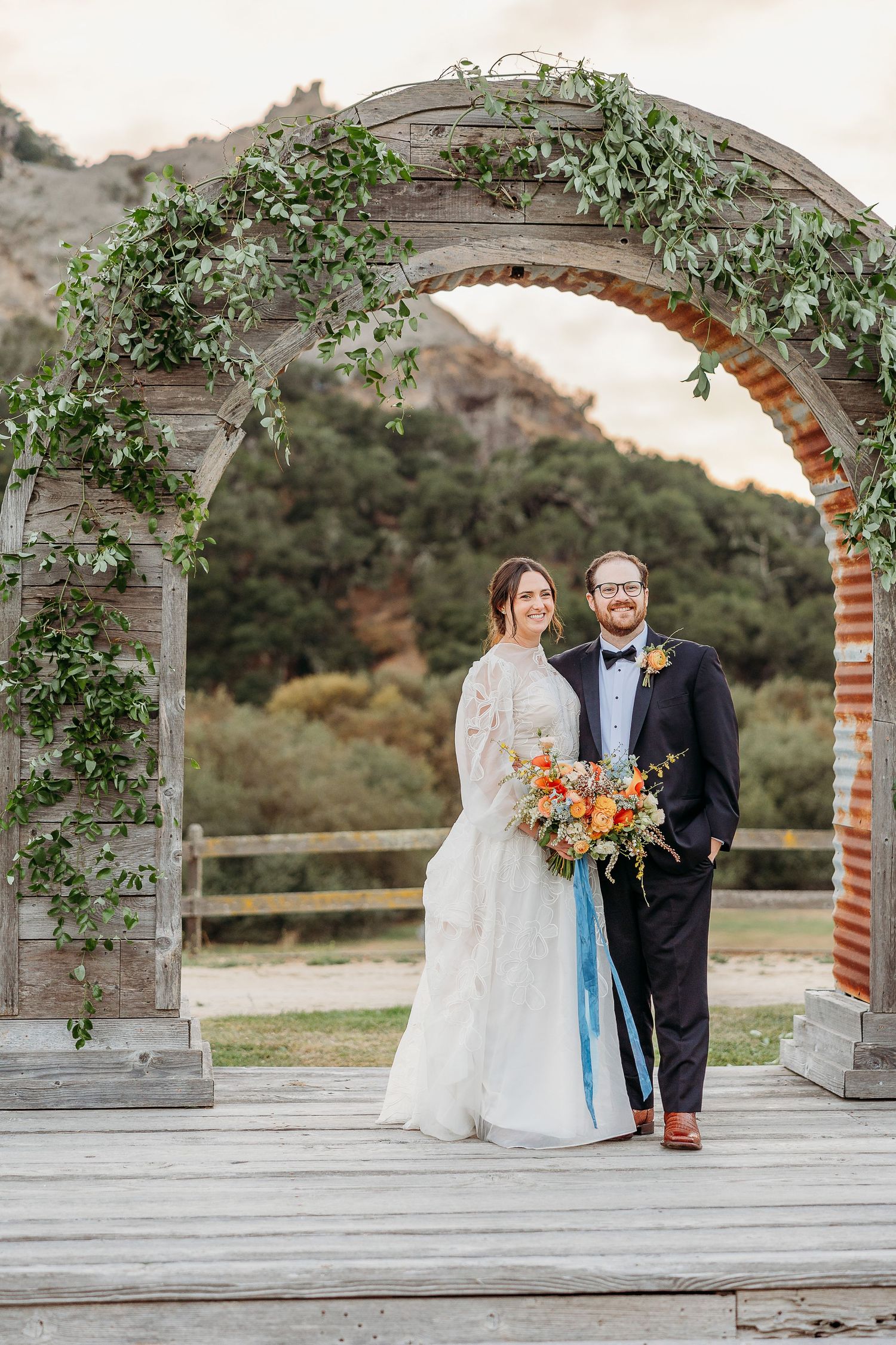 A couple poses beneath a vine-covered wedding arch with an orange bouquet and teal ribbon details.