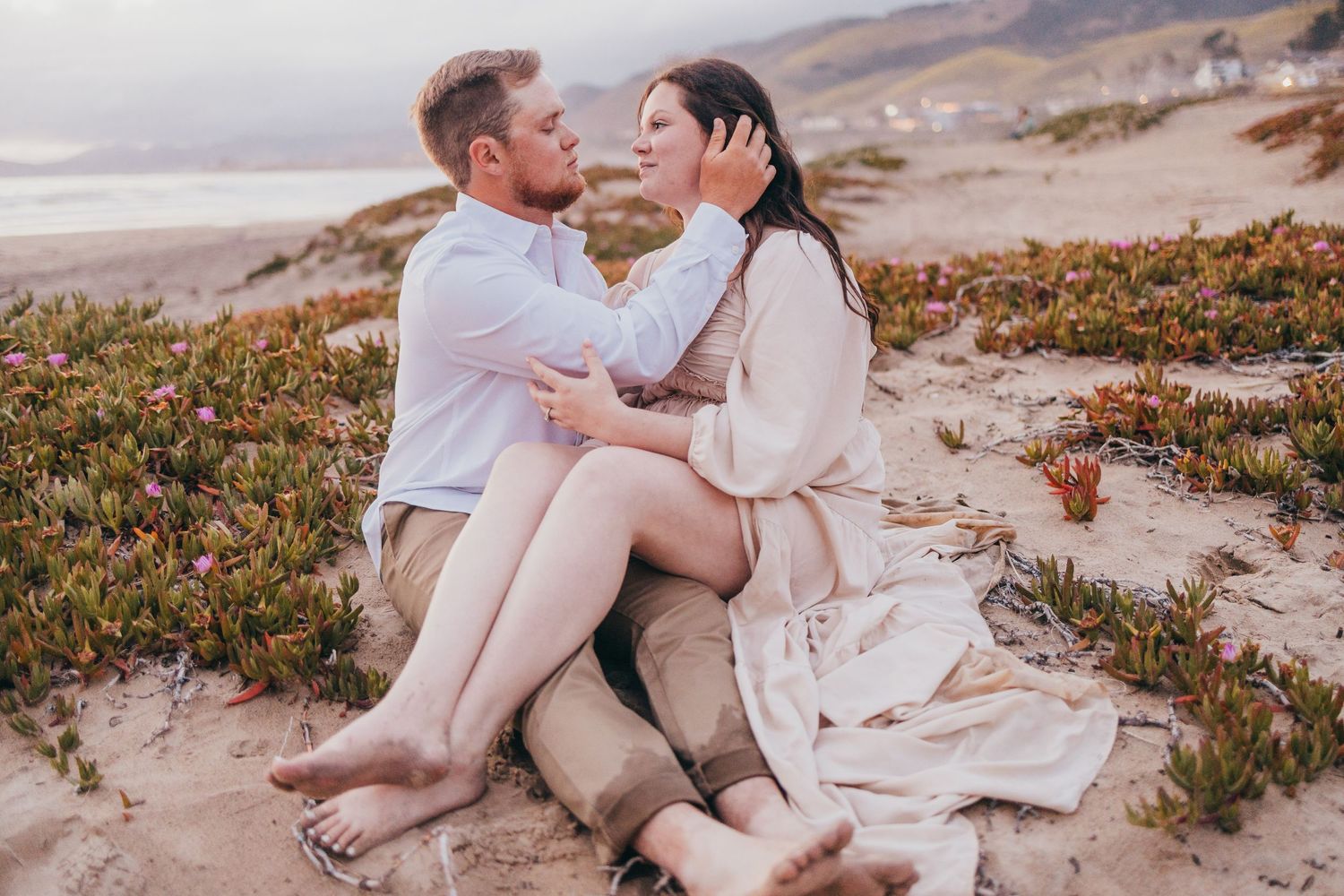 Couple shares tender moment while sitting on coastal dunes with wildflowers.