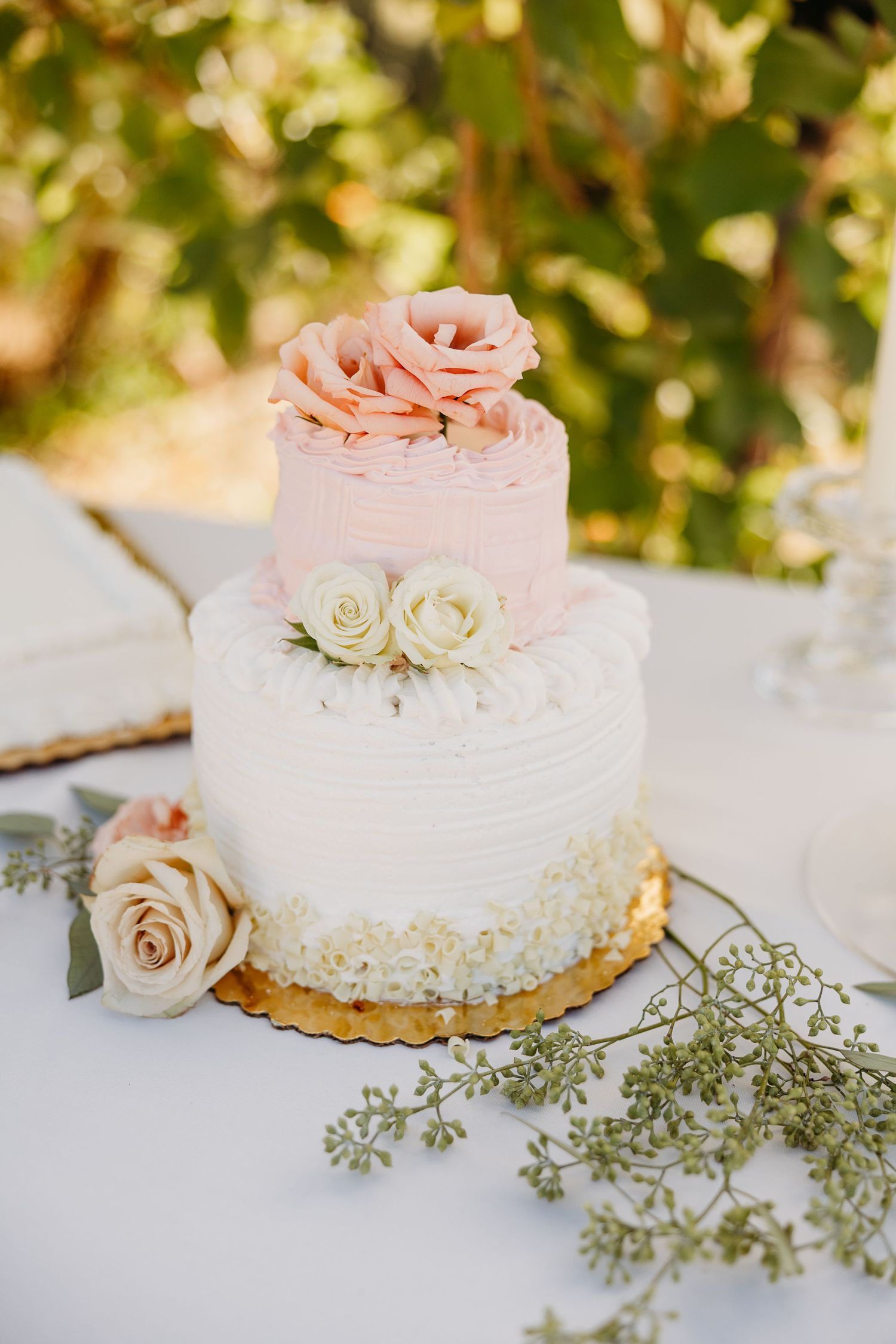 A two-tier wedding cake decorated with pink and white roses on a white tablecloth with delicate greenery.