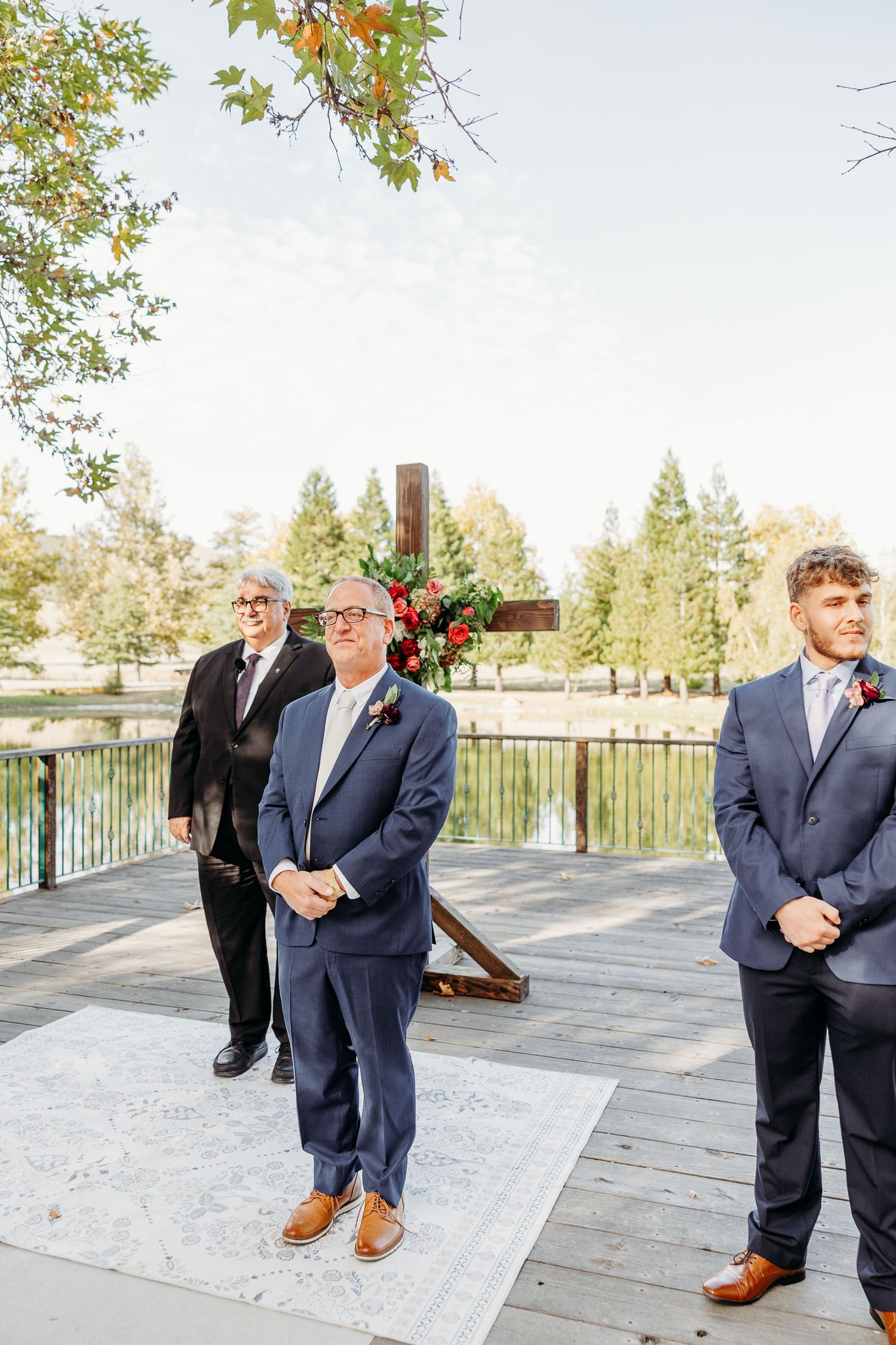 Groomsmen in navy suits and brown shoes standing on a deck during an outdoor wedding ceremony.