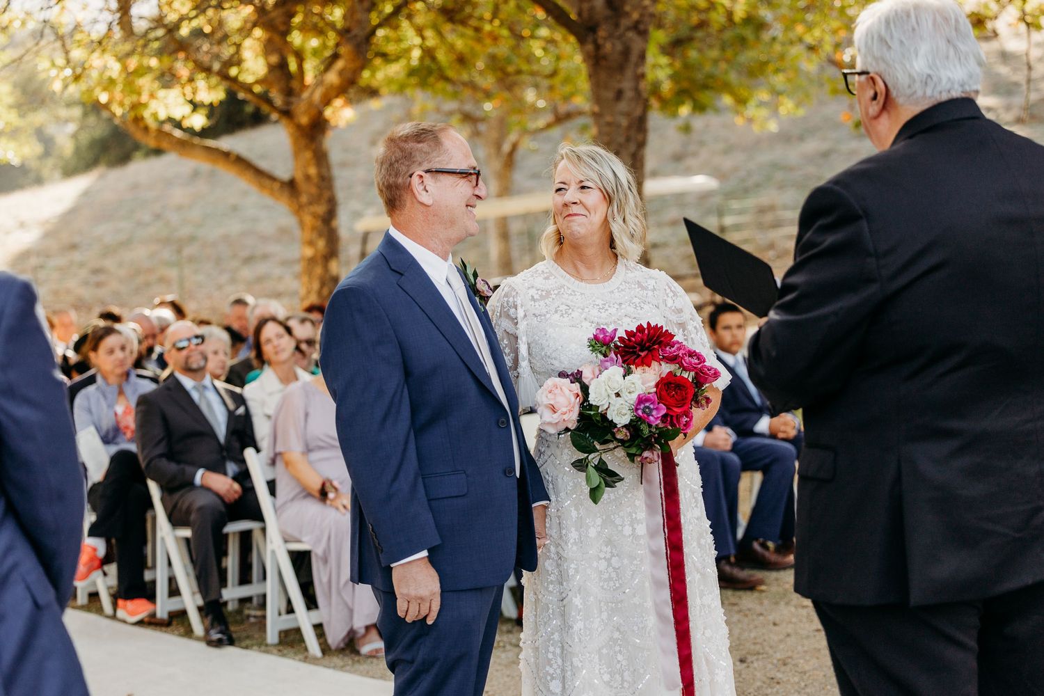 Outdoor wedding ceremony setting with guests gathered under autumn trees.