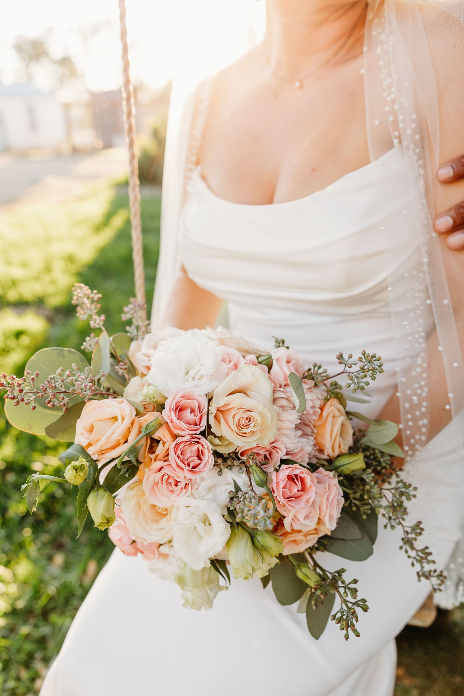 Elegant floral wedding bouquet with peach roses, white flowers and greenery against a white dress.