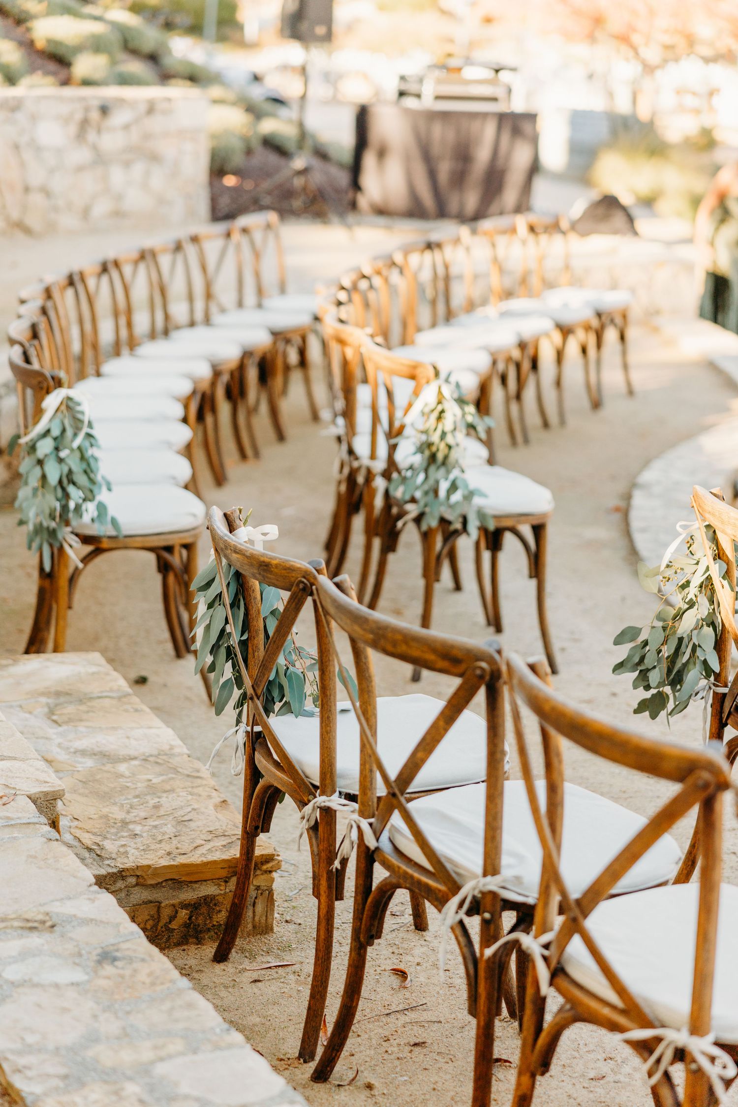 Wooden crossback chairs decorated with greenery line up for outdoor wedding ceremony.
