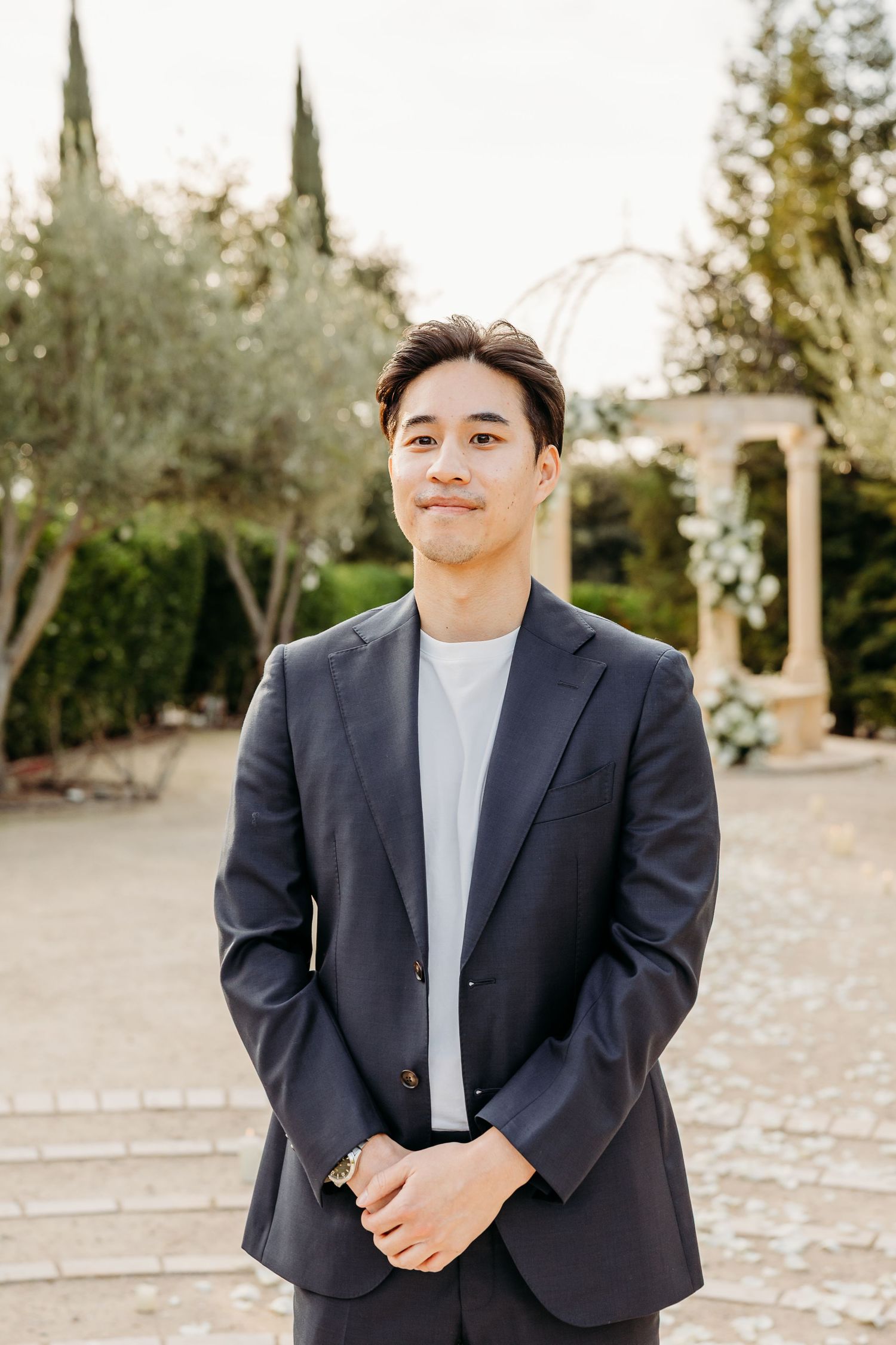A person in a navy suit jacket and white t-shirt stands in a garden setting with stone columns and cypress trees right before he proposes