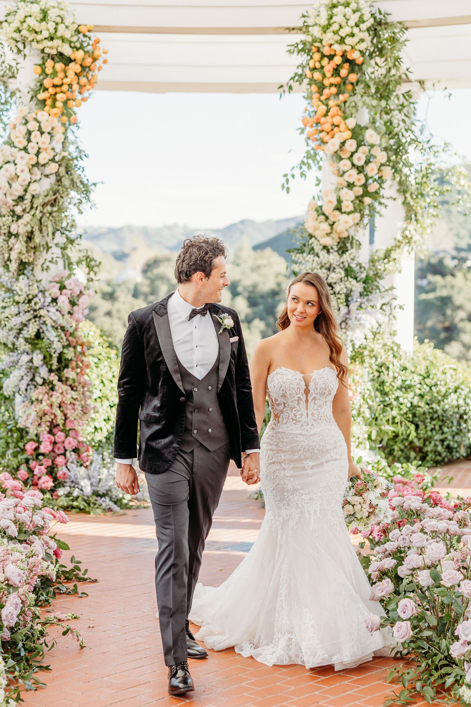 Wedding couple walking under floral arch with mountain views and romantic garden decor.