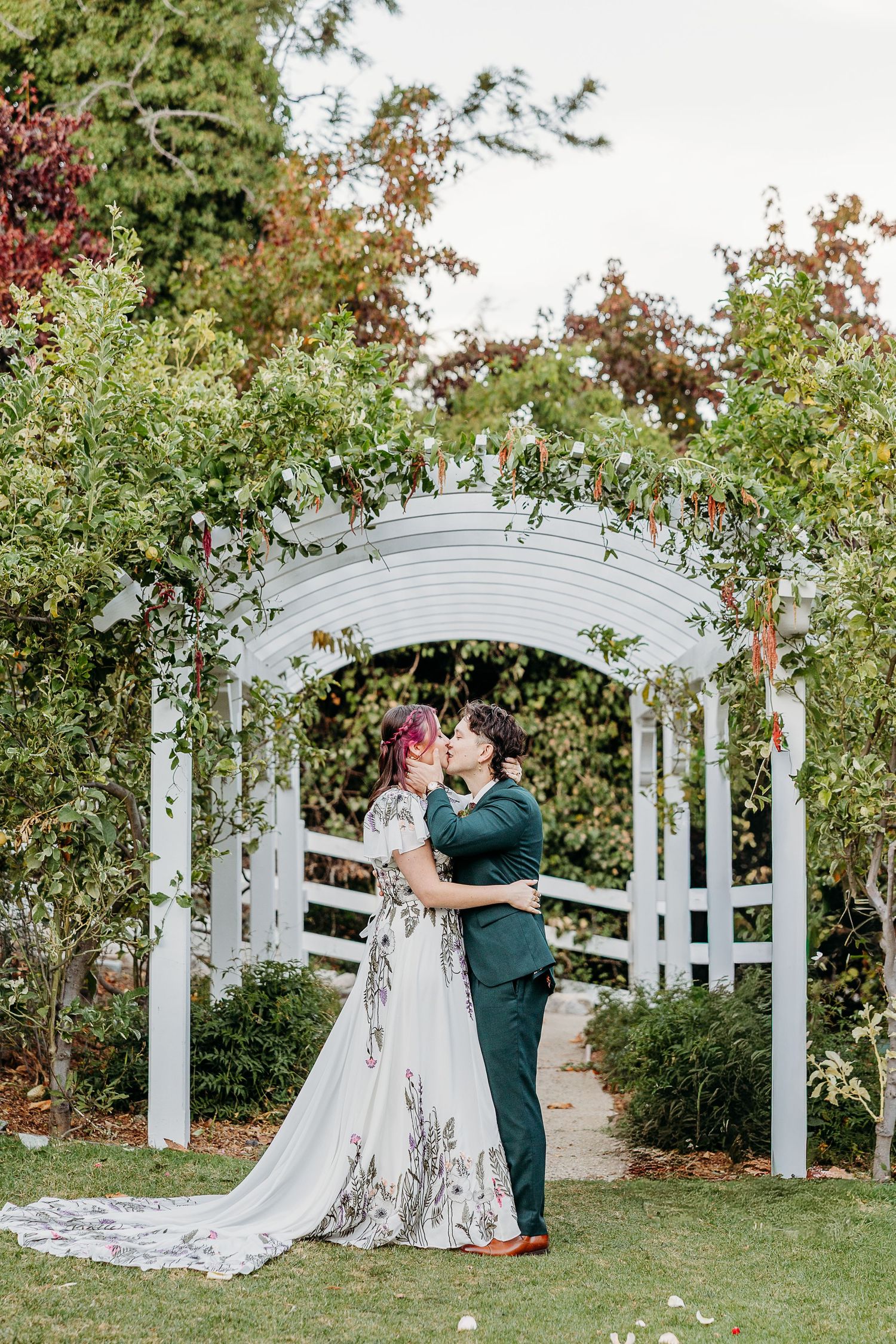 Couple shares intimate moment under white garden arbor during wedding ceremony.
