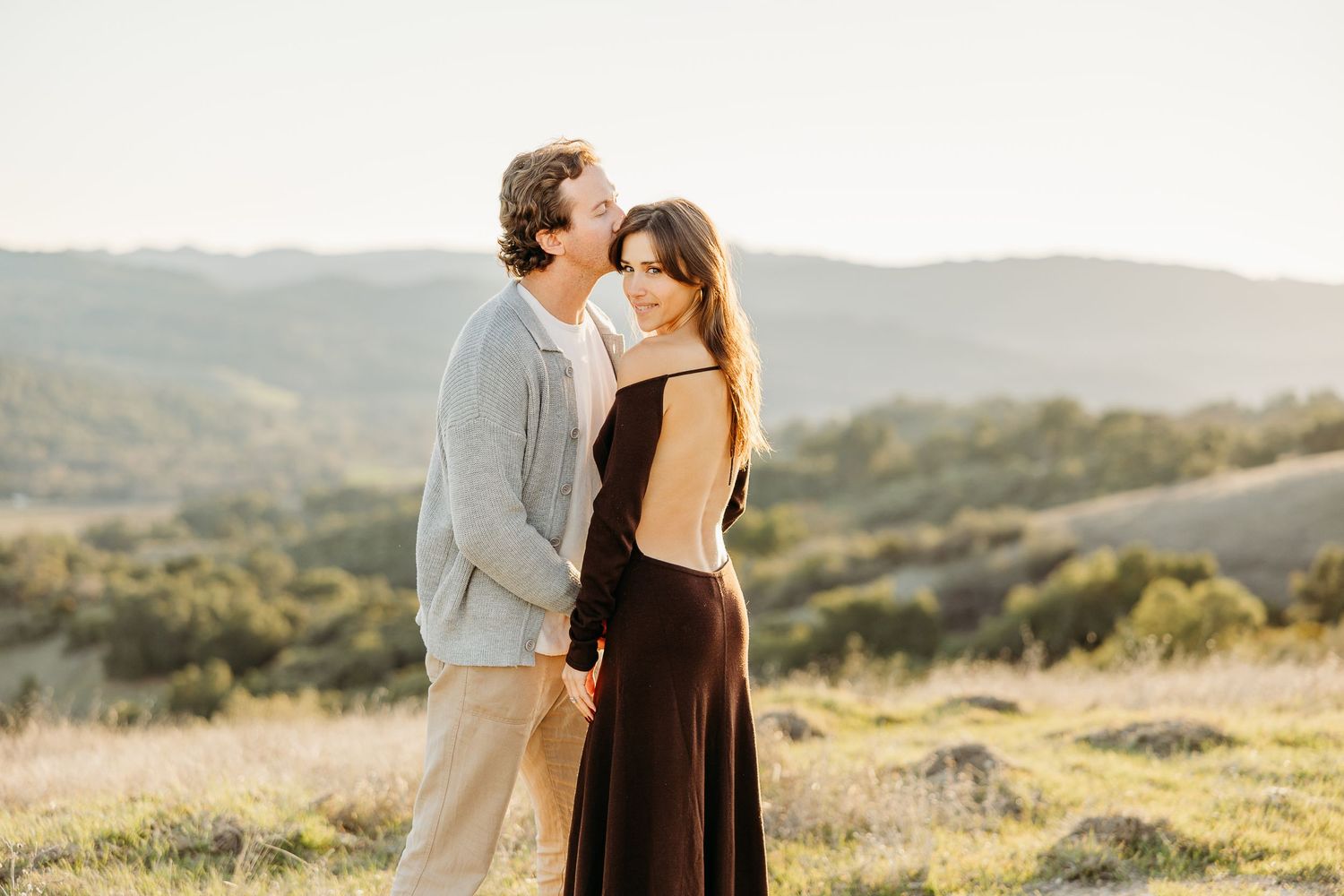 A couple embraces on a hilltop during golden hour with mountains and rolling hills in the background.
