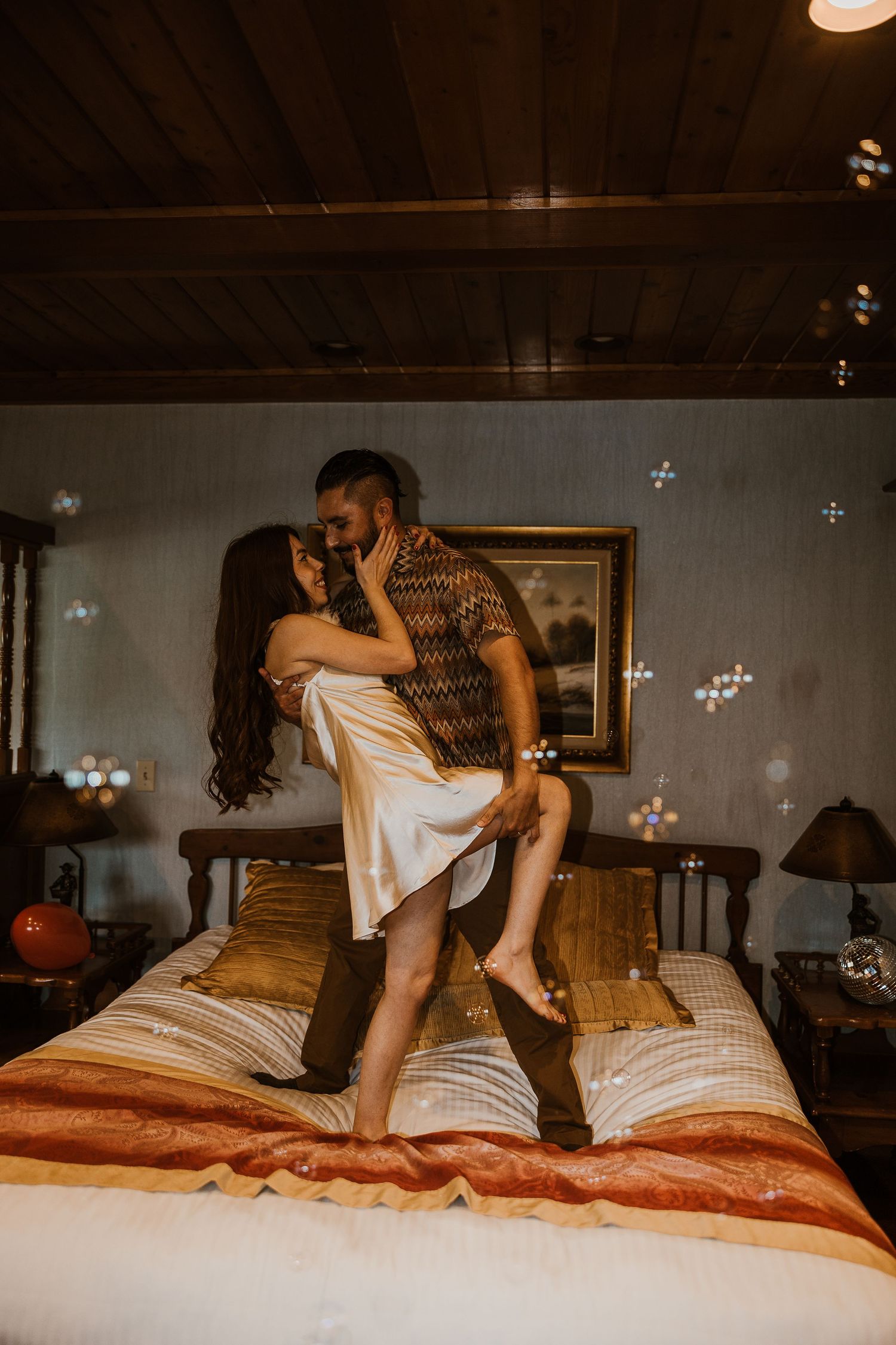 Romantic couple sharing an intimate moment on a warm-toned bed with string lights and a cozy bedroom atmosphere.