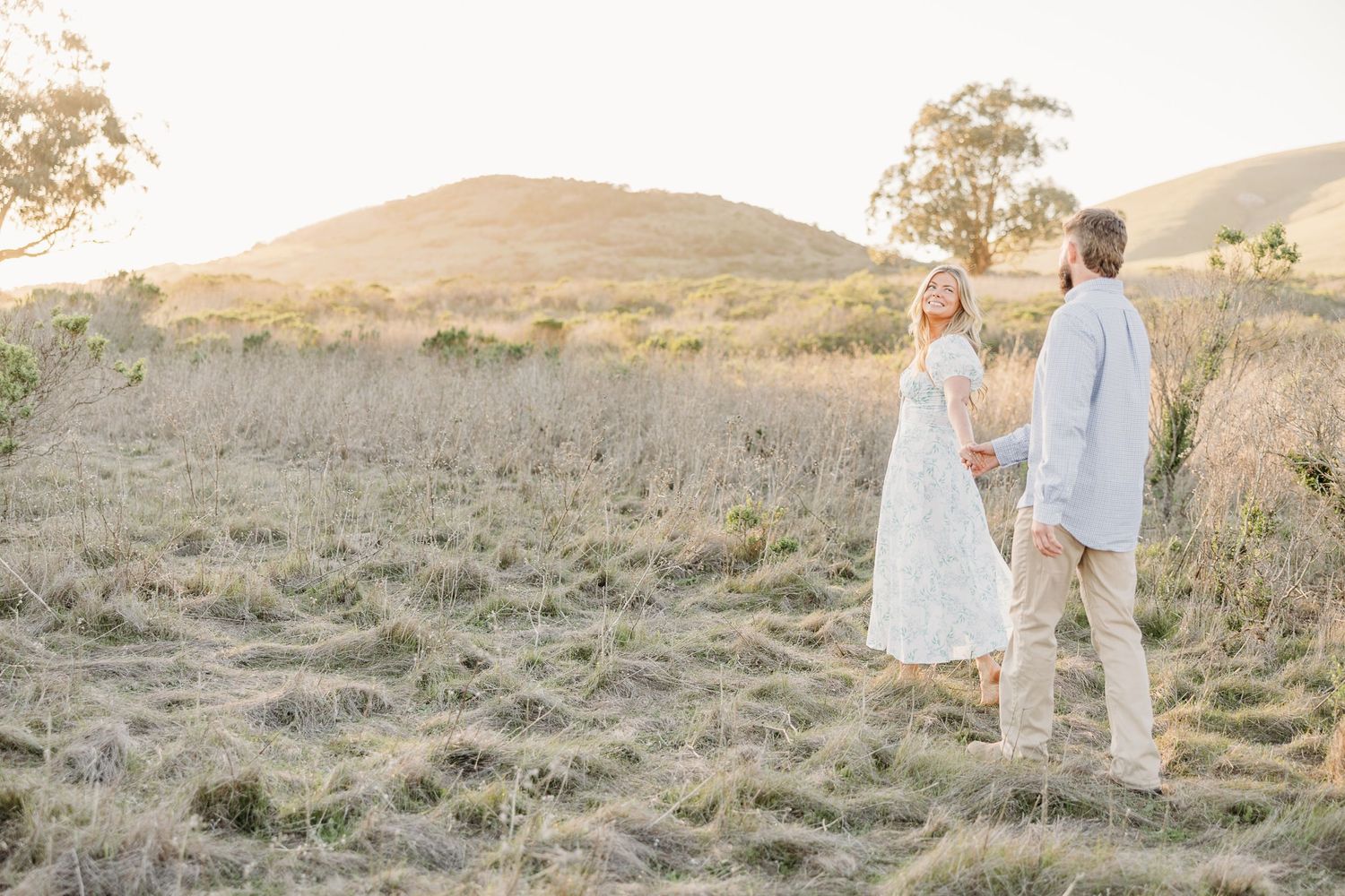 A couple shares a romantic moment at sunset in a golden field with rolling hills in the background.