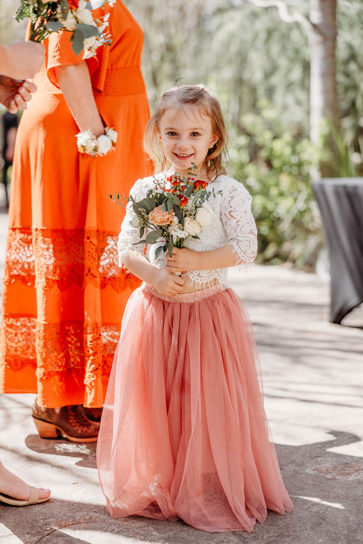Young attendant in pink tulle skirt holding floral bouquet at outdoor wedding.