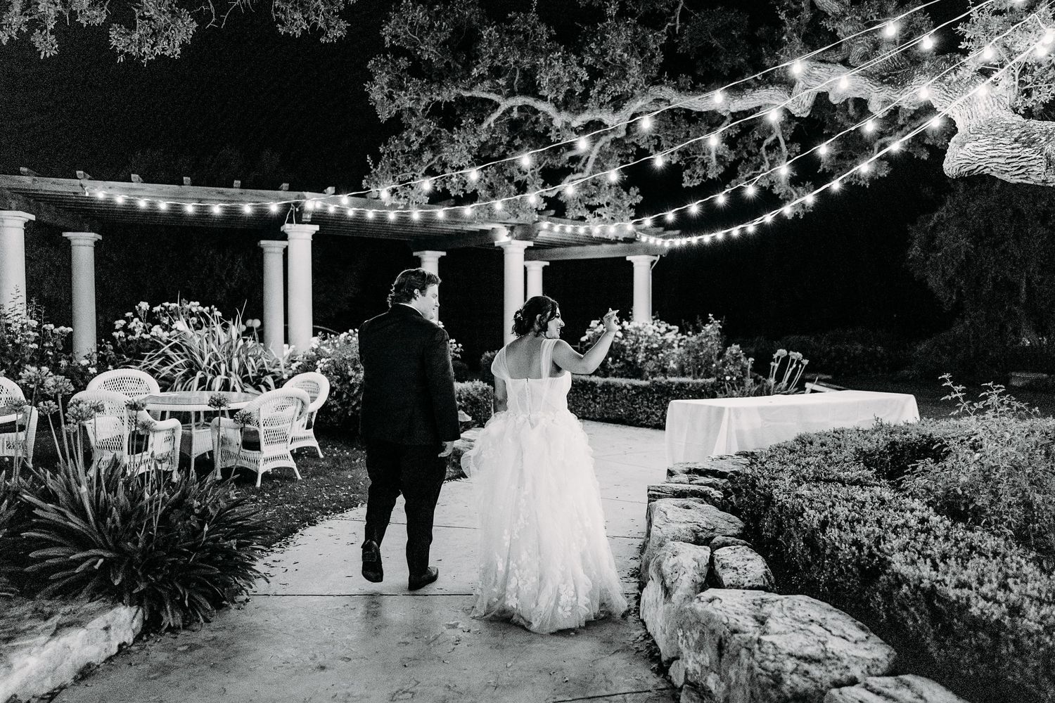 Silhouette of couple walking along garden path under string lights at evening wedding.
