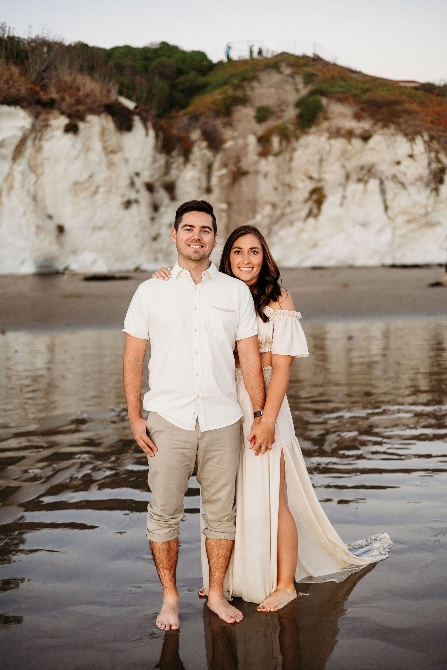 A couple poses together on a beach with dramatic white cliffs in the background during sunset.