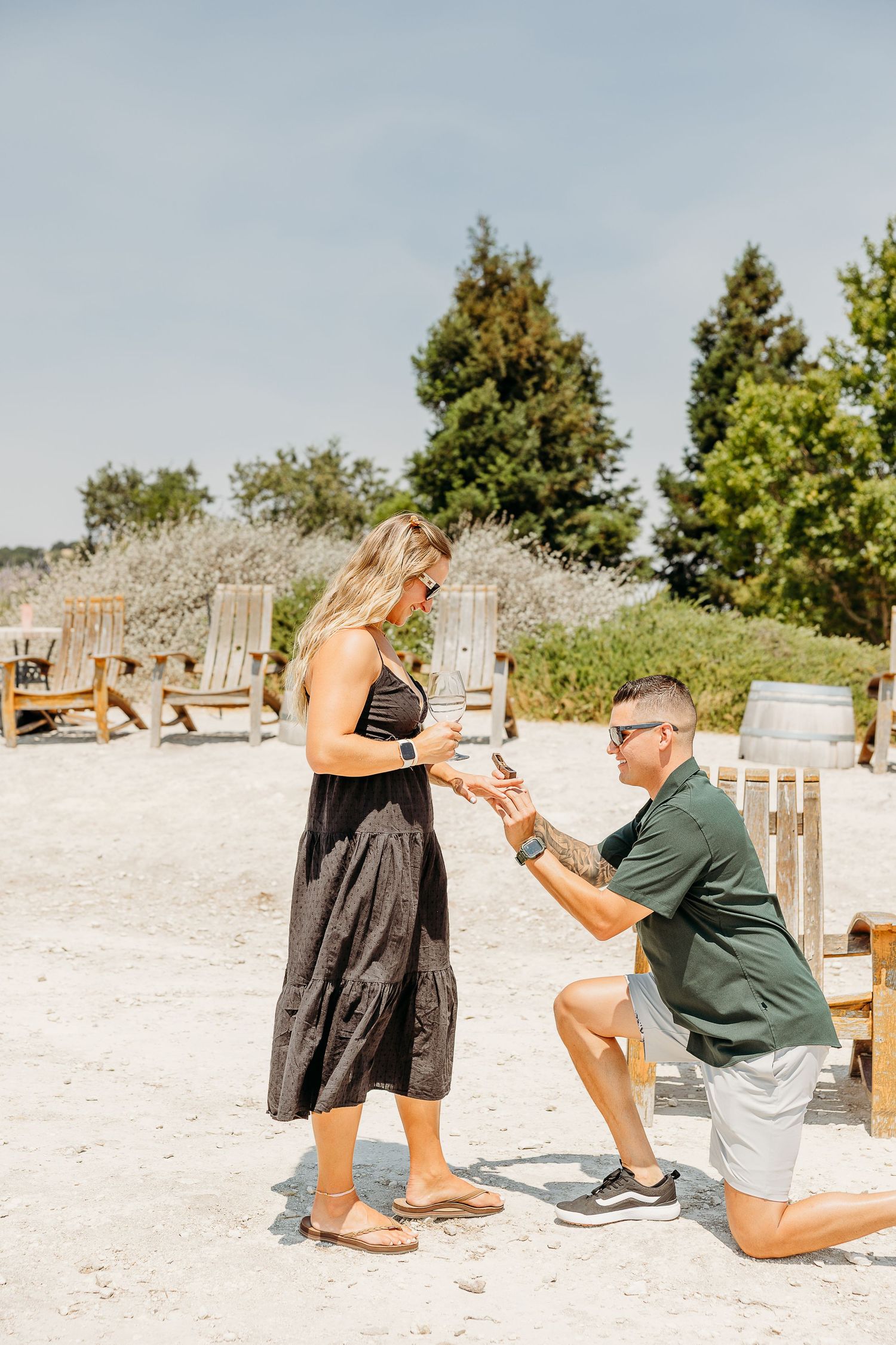 A couple shares a romantic proposal moment on a sandy beach with trees in the background.