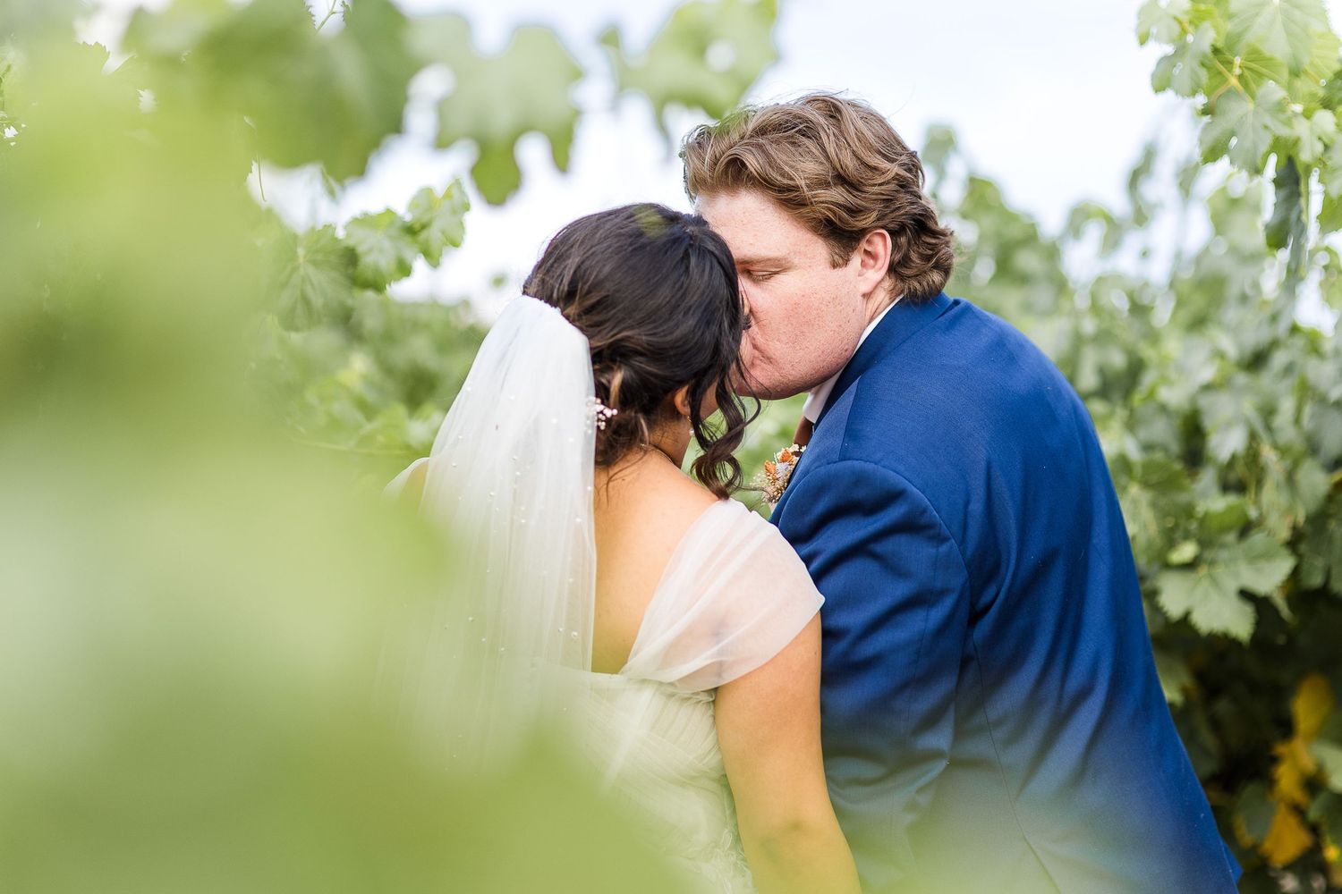 Intimate wedding kiss captured through vineyard leaves on sunny day.