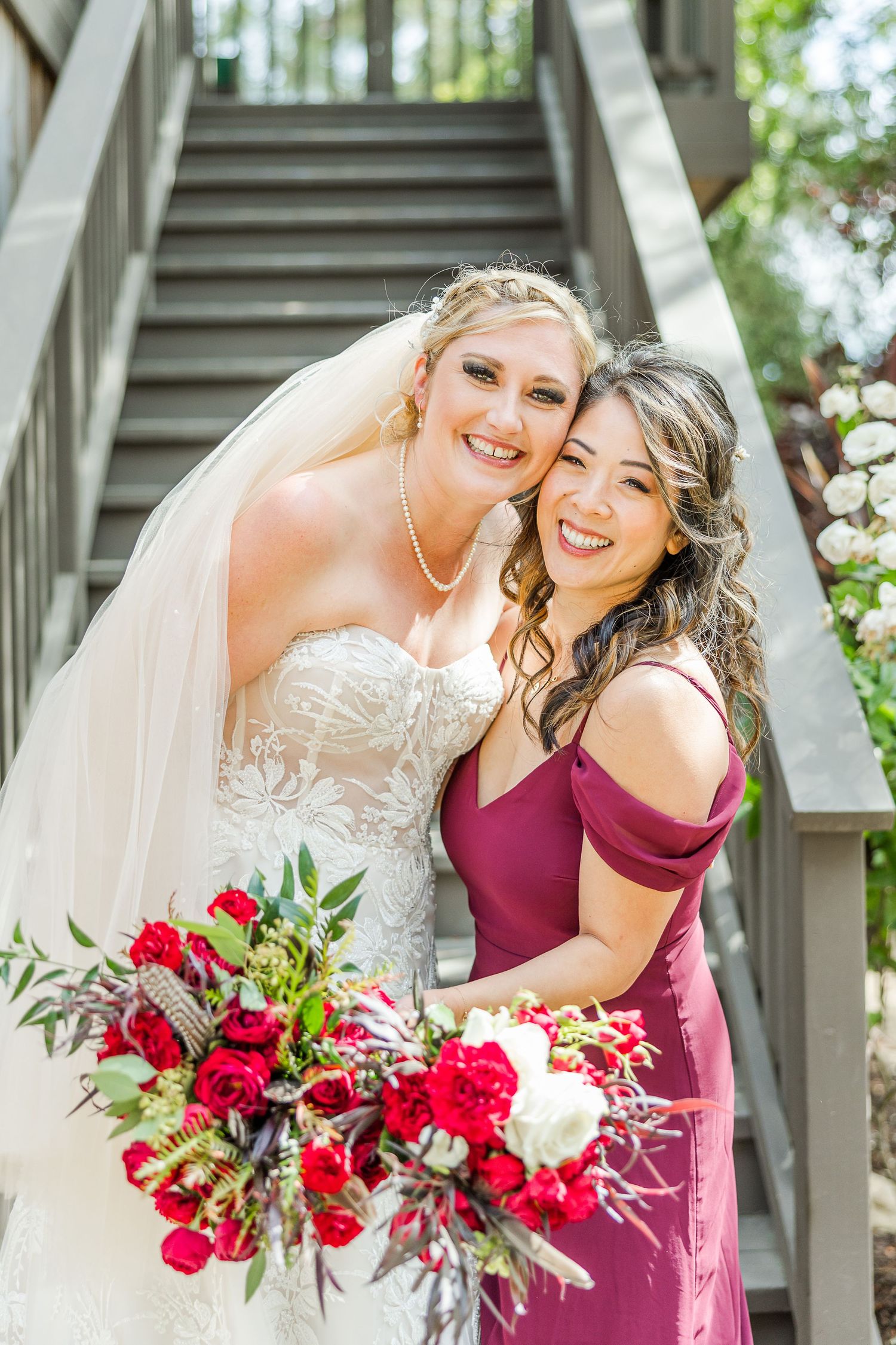 Two people pose together on outdoor stairs surrounded by greenery with a vibrant red bouquet.