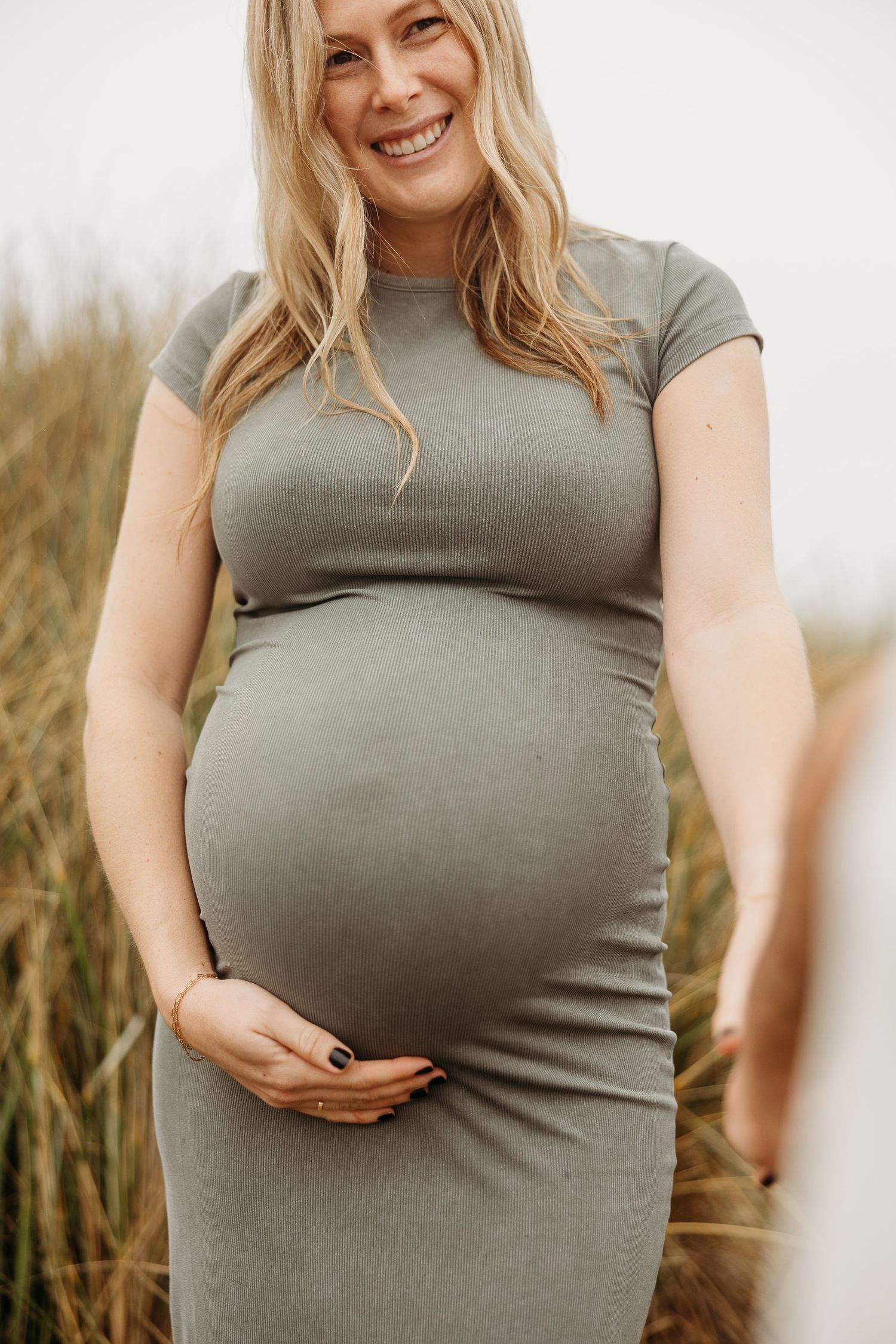 Maternity photo shows woman in fitted sage green dress cradling her baby bump in outdoor setting.