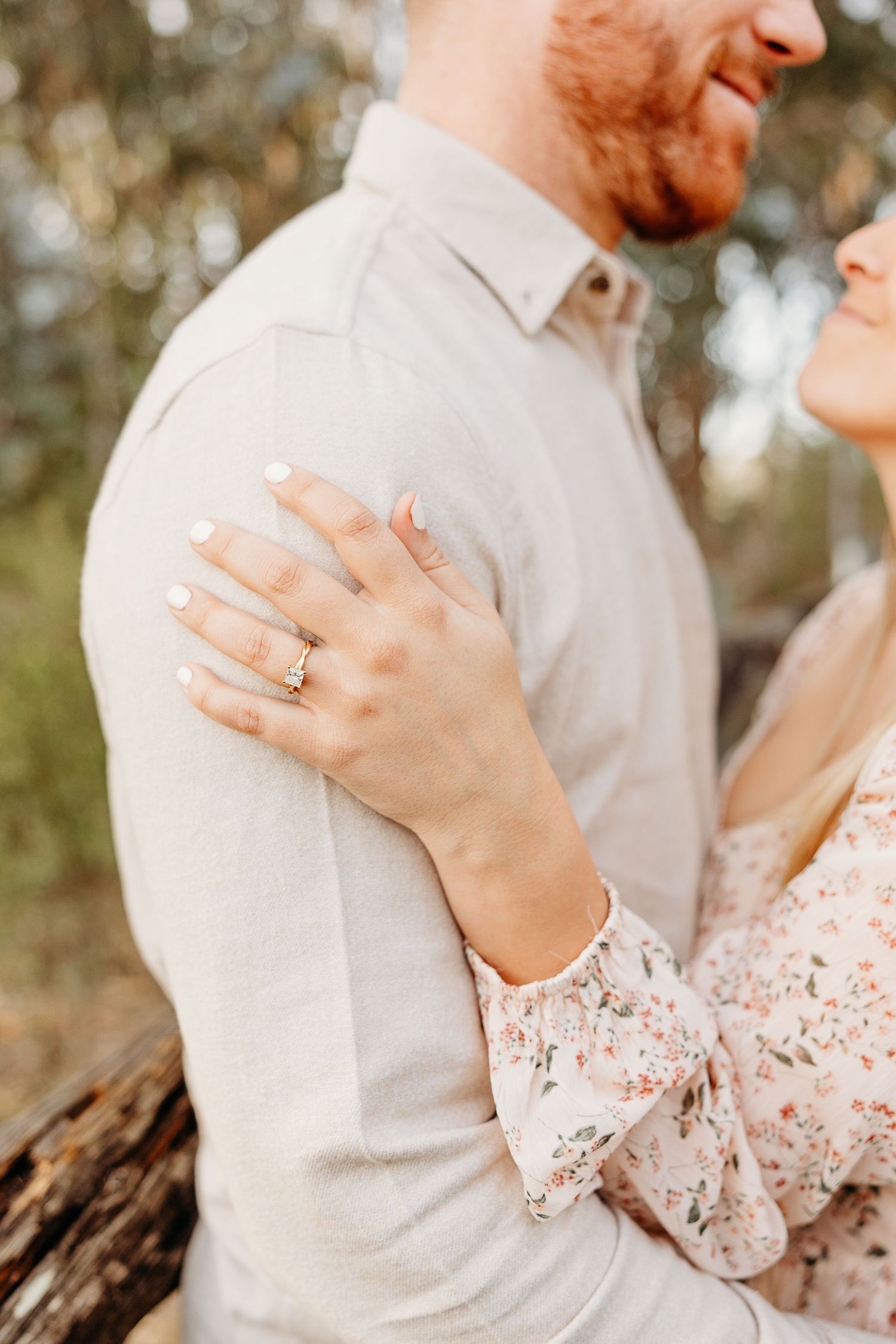 Close up of engagement ring as couple embraces in soft natural lighting.