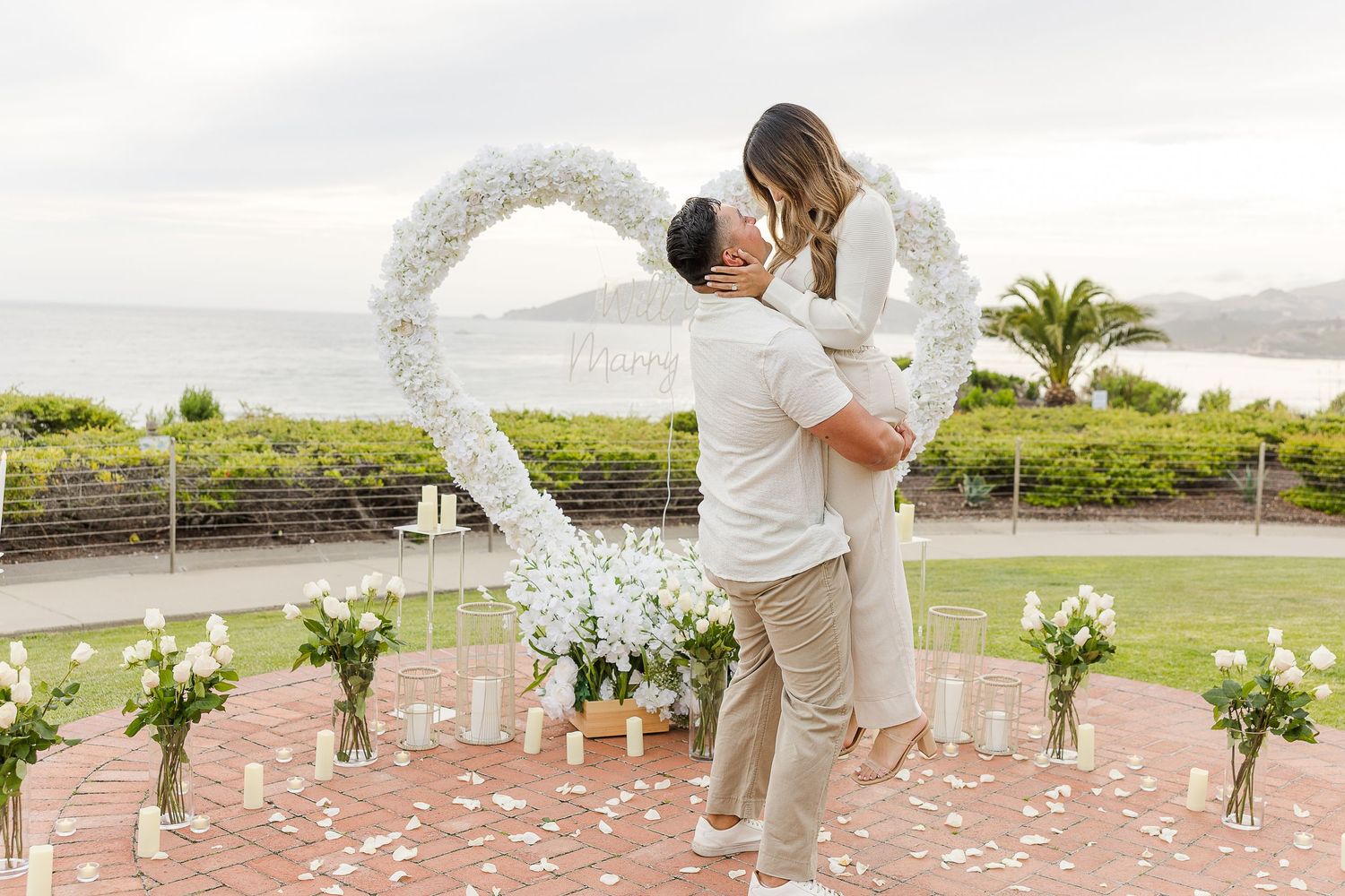 Wedding ceremony setup with white floral arch and rose petals scattered along a stone pathway.