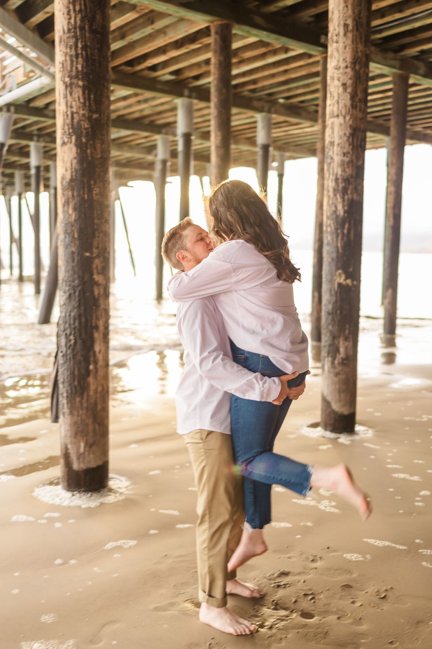 Couple embracing under wooden pier on beach during romantic photoshoot.