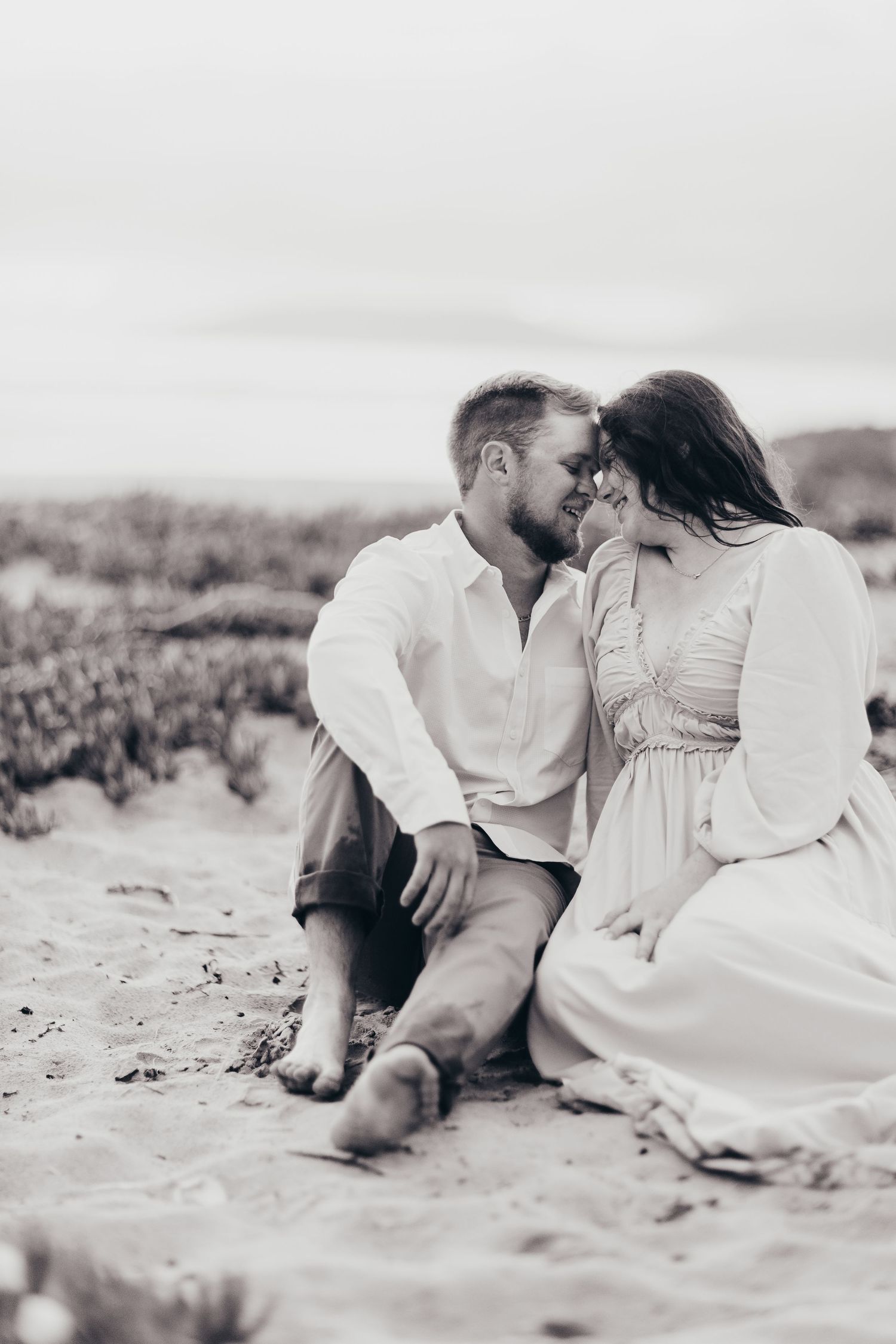 Black and white portrait of a couple sitting intimately on coastal dunes.