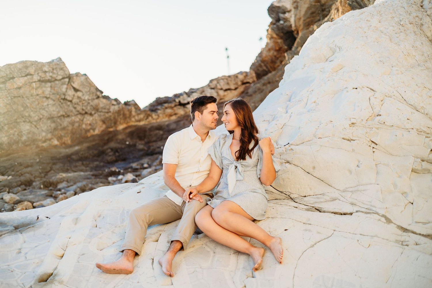 A couple shares a joyful moment while sitting on coastal rocks during golden hour.