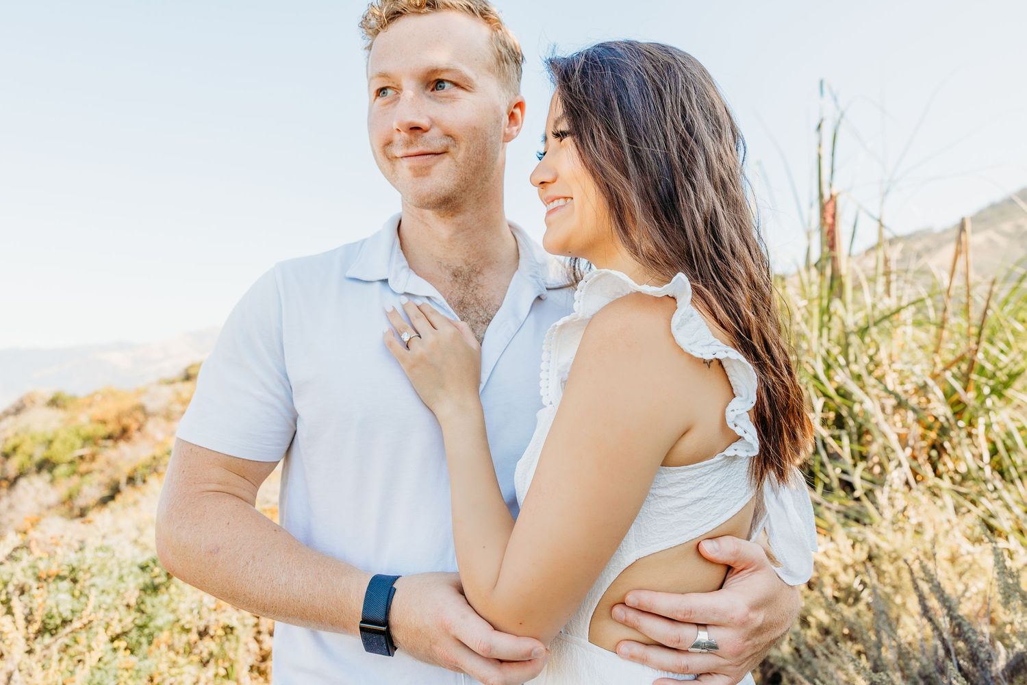 Couple embracing in white outfits against a sunny mountain landscape backdrop.