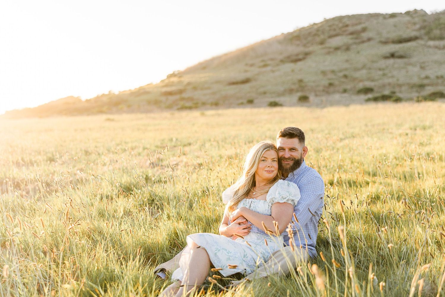 A couple sits together in golden field at sunset, embracing on a grassy hillside with warm sunlight glowing behind them.