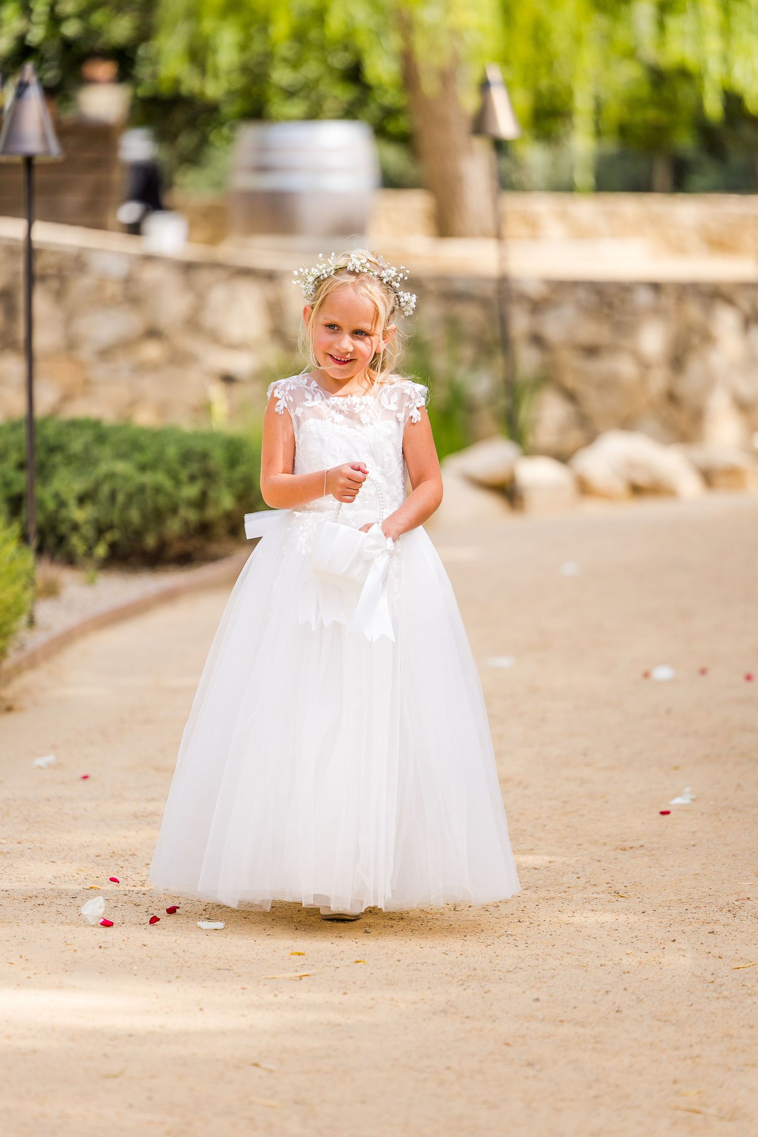 A flower girl in a white tulle dress walks down a garden path.