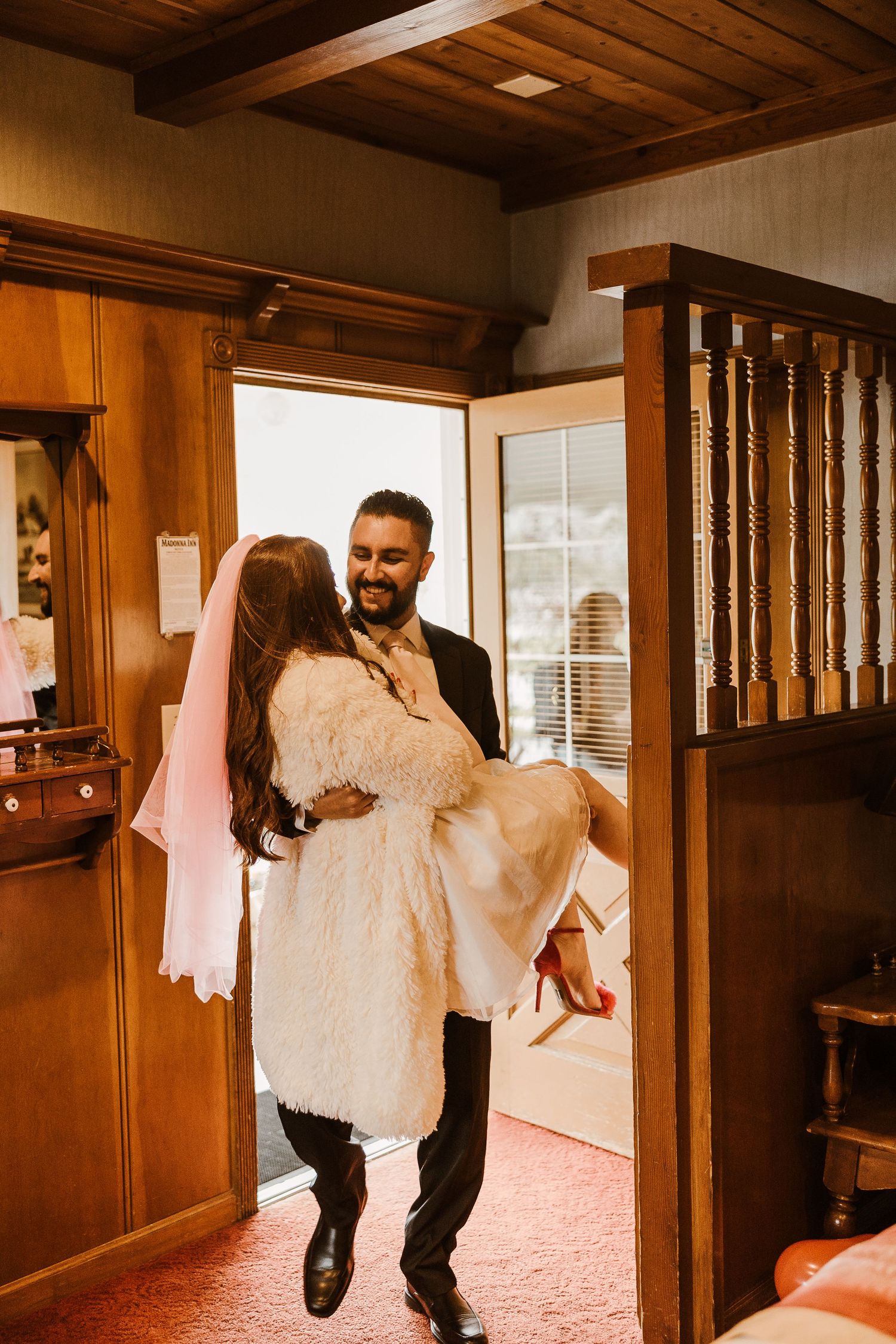 A couple in a rustic wooden interior with the bride wearing white being carried across the threshold in romantic lighting.